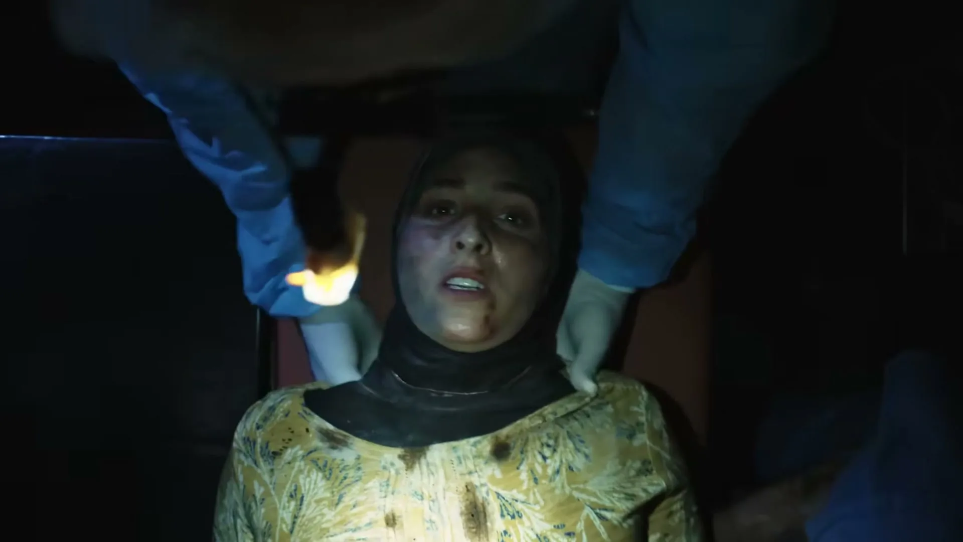 A woman in a hijab lies on a gurney, looking up with a pained expression, as medical staff in blue gloves attend to her in a dark, emergency setting.