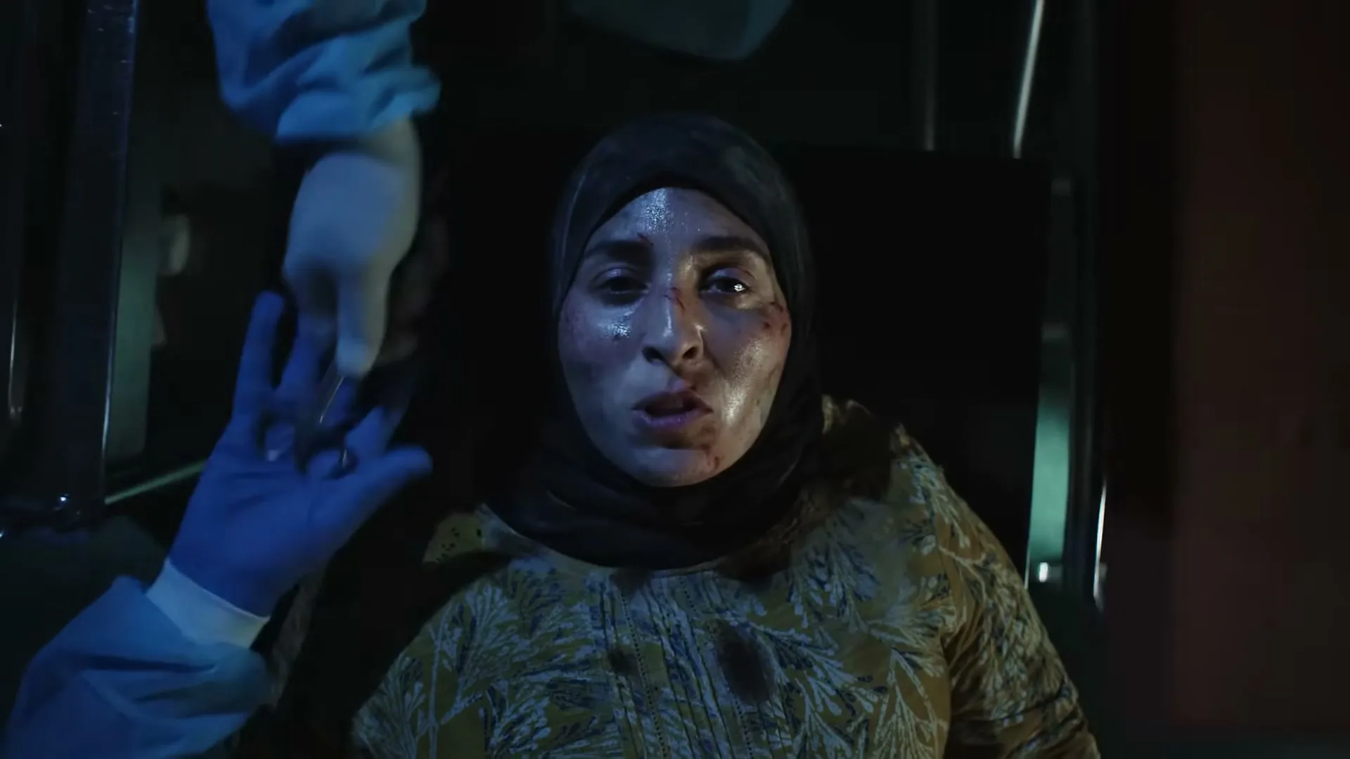 A woman with a headscarf and blood on her face lies on a stretcher, looking up in distress as medical personnel attend to her in a dark, chaotic setting.