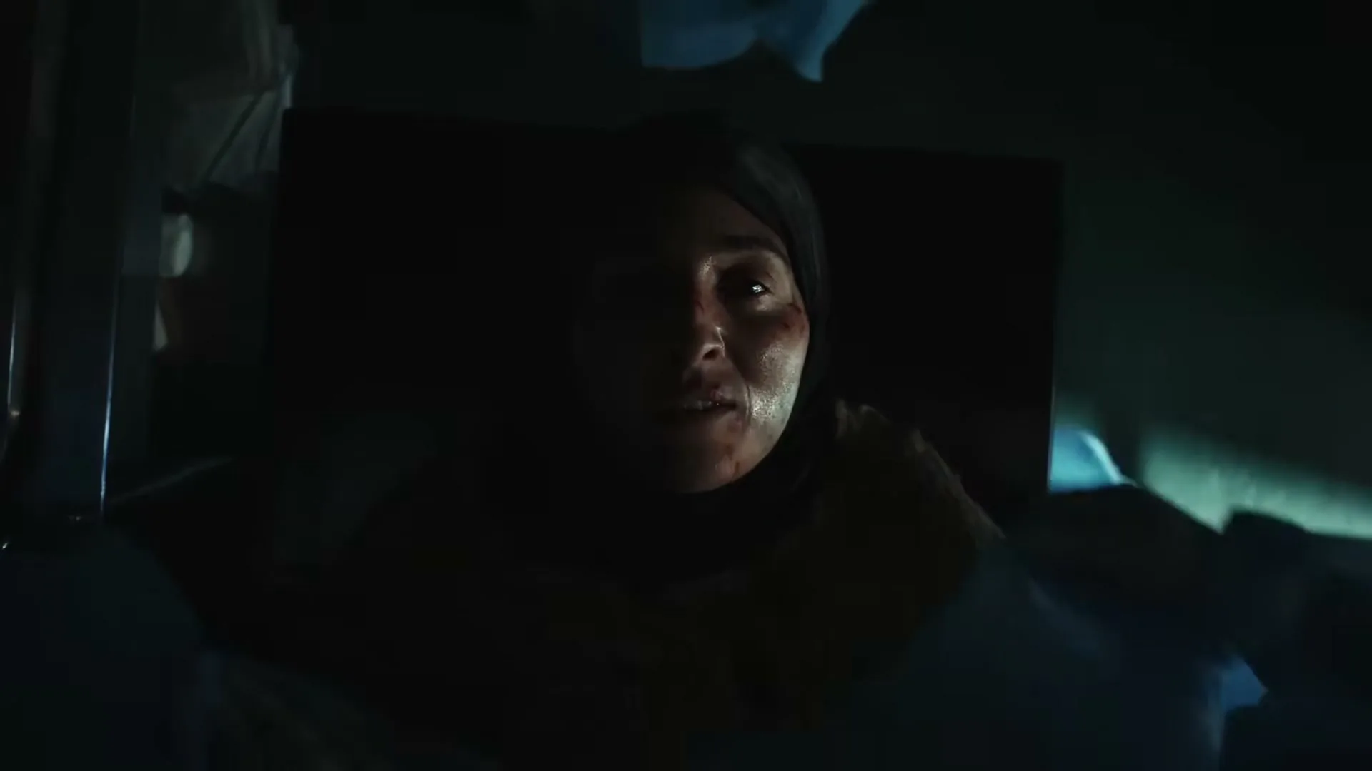 A woman with a headscarf lies on a gurney, looking up with a pained expression in a dark, medical setting.