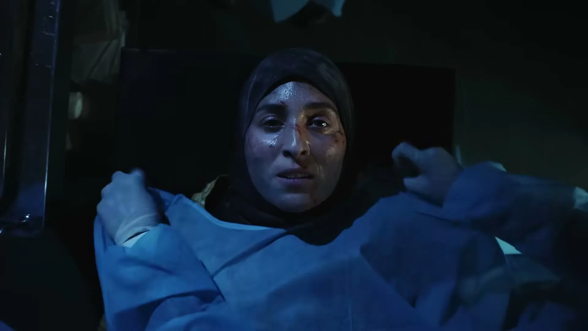 A woman in a hijab and medical gown, with blood on her face, looks up with a pained expression in a dark, chaotic interior.