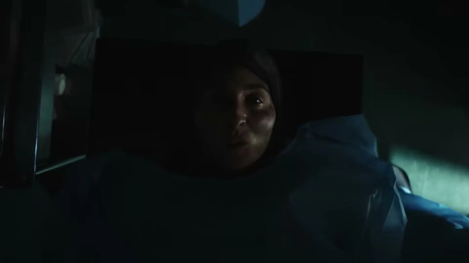 A woman with a head covering and a blue medical gown lies on a gurney, looking up with a pained expression in a dark, confined space.
