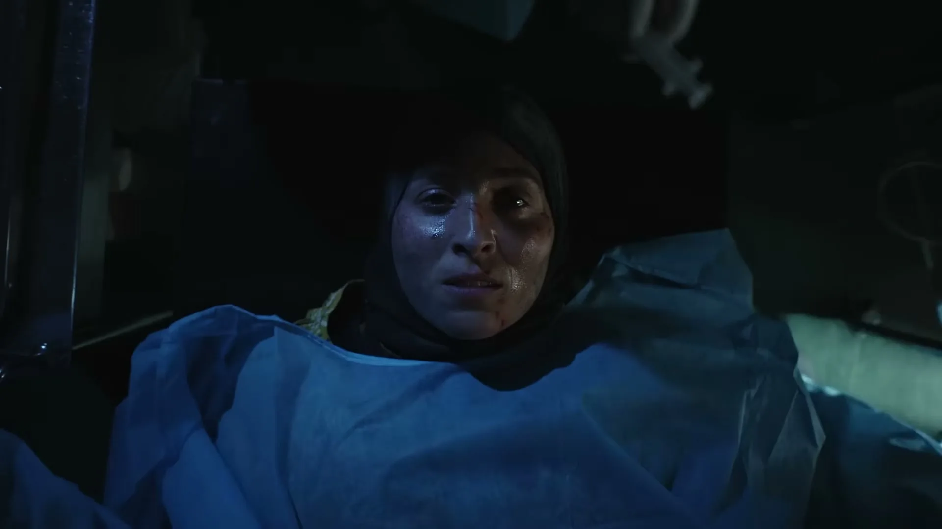 A woman with a headscarf and a blue medical gown lies on a gurney, looking up with a pained expression in a dimly lit medical setting.