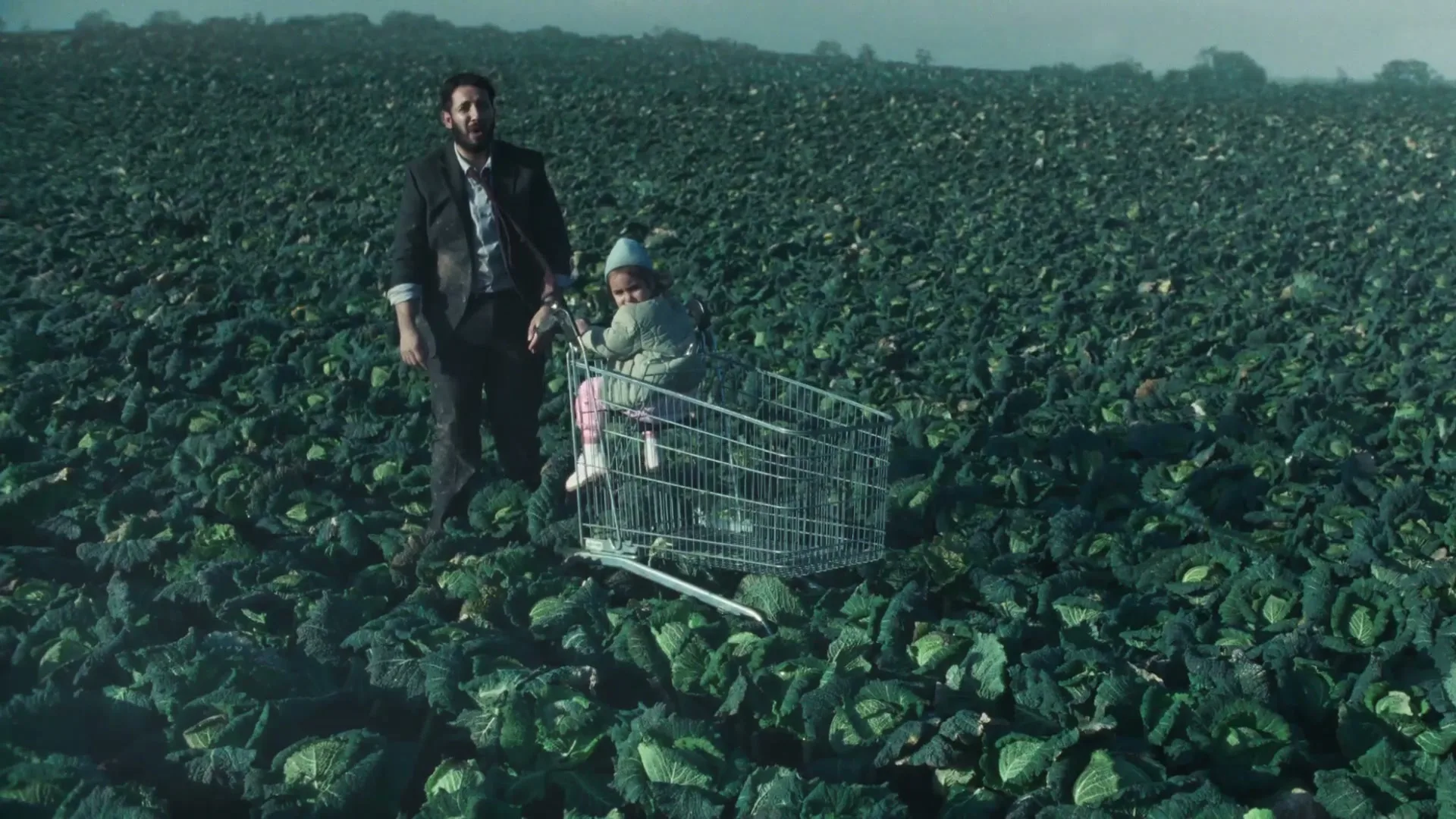 A man in a suit stands next to a child in a shopping cart in a vast field of green cabbage under a hazy sky.
