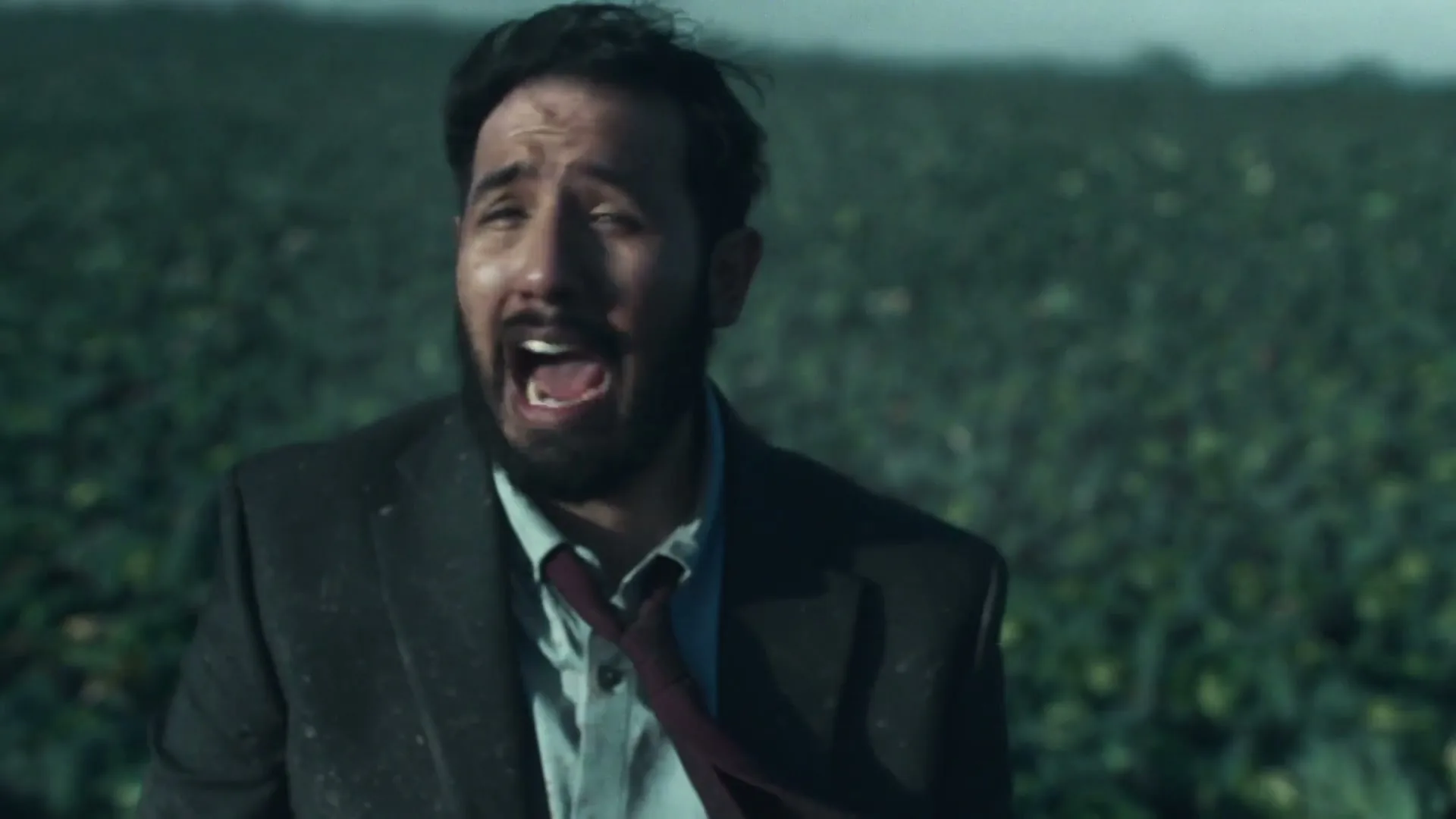 A man with a beard and a dirty suit looks around frantically in a field of green leafy plants, appearing lost and distressed.