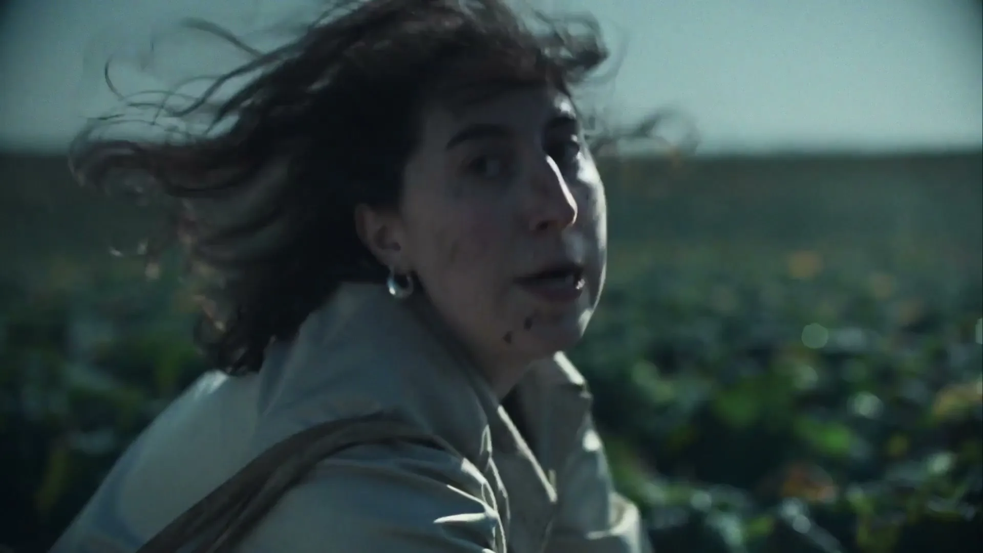 A woman with dark hair and a light coat runs through a field of green plants, looking distressed with dirt on her face.