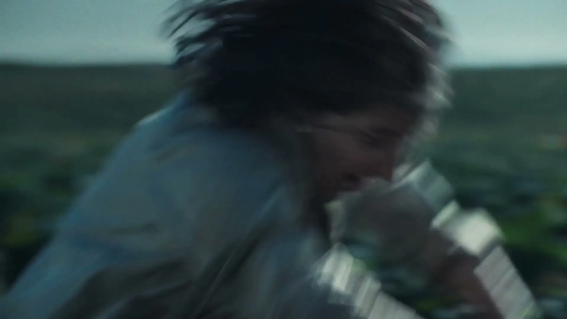 A person with long dark hair and a light shirt is seen in motion, with a blurred background of green plants under a grey sky.