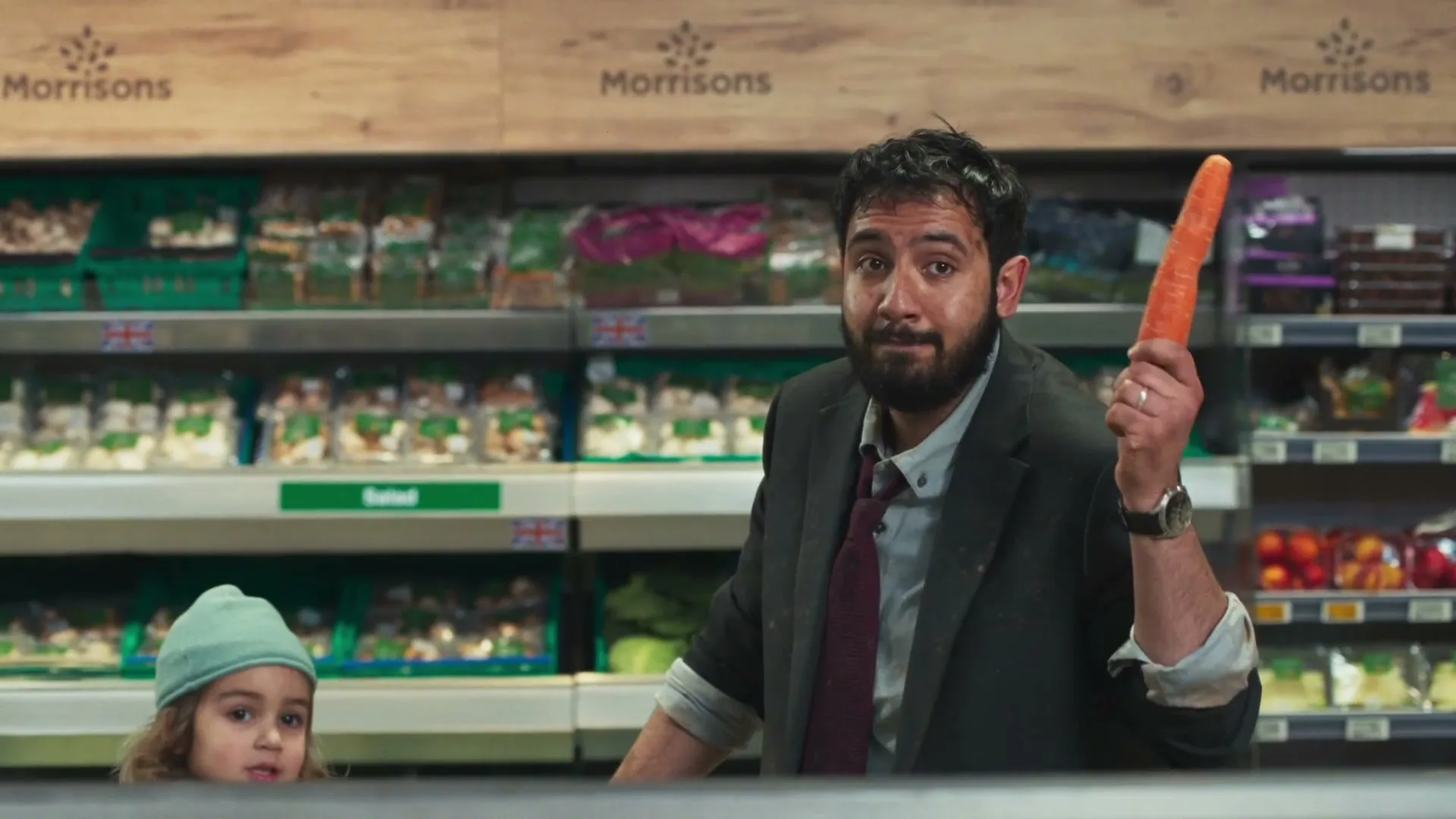 A man and a child pop up from behind a counter in a supermarket, holding carrots and looking surprised.