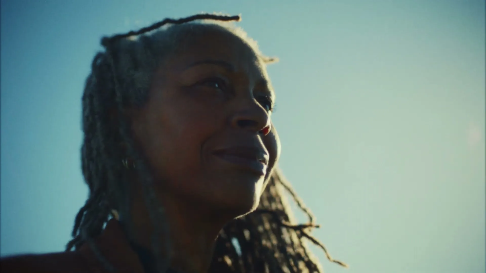 A woman with dreadlocks smiles, looking up and to the right, with a bright blue sky in the background.