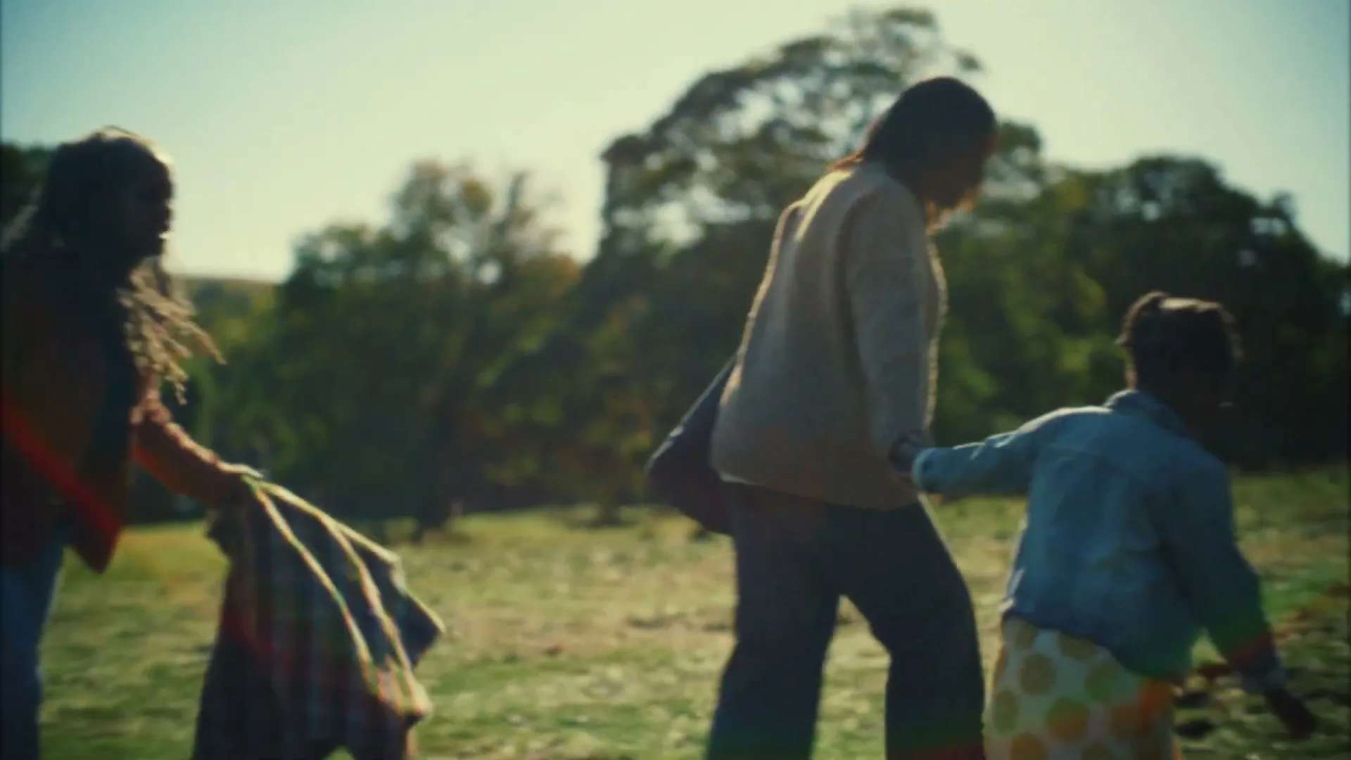 A family of three, two adults and a child, walk across a grassy field on a sunny day, holding hands.