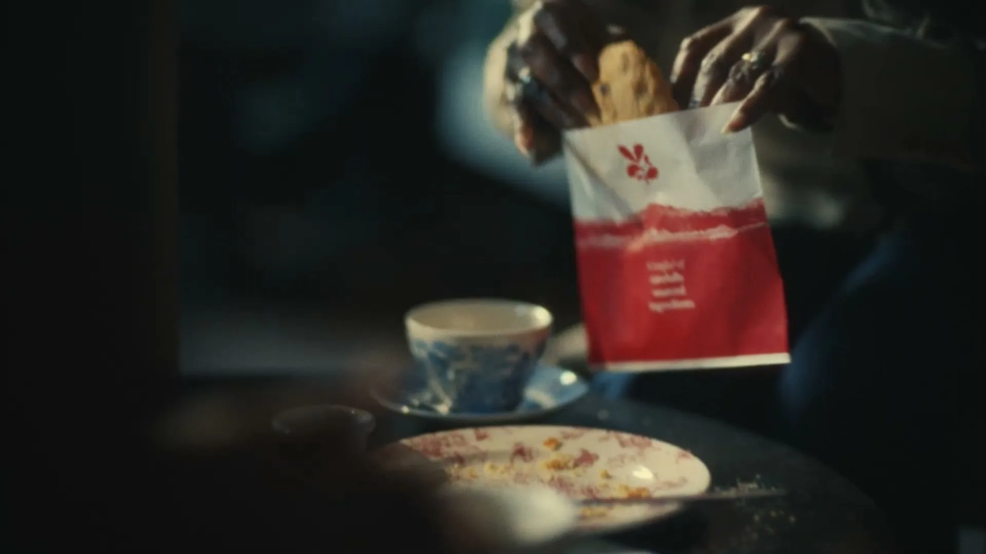 A person's hands take a cookie from a white and red paper bag on a table with teacups and a plate with crumbs, in a dimly lit interior.
