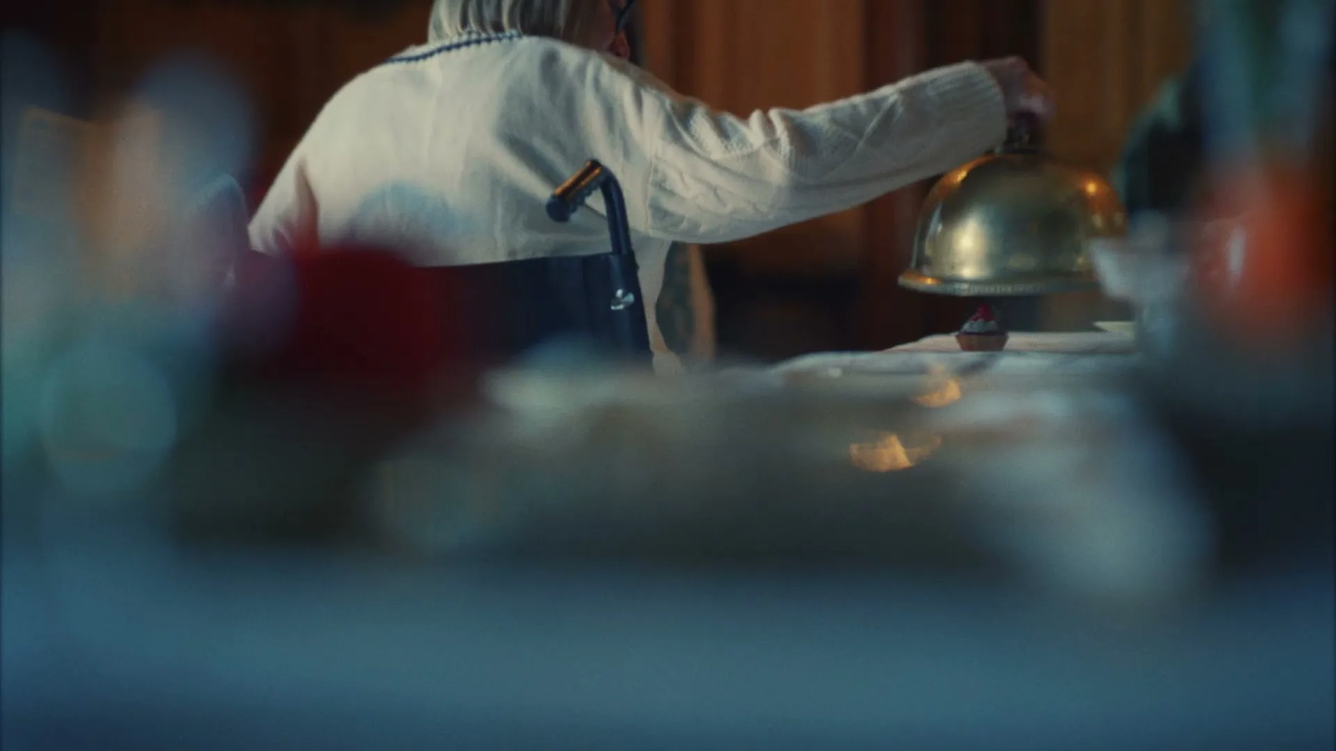 An older woman in a wheelchair, wearing a white sweater, lifts a golden cloche from a small dessert on a table in an interior setting.