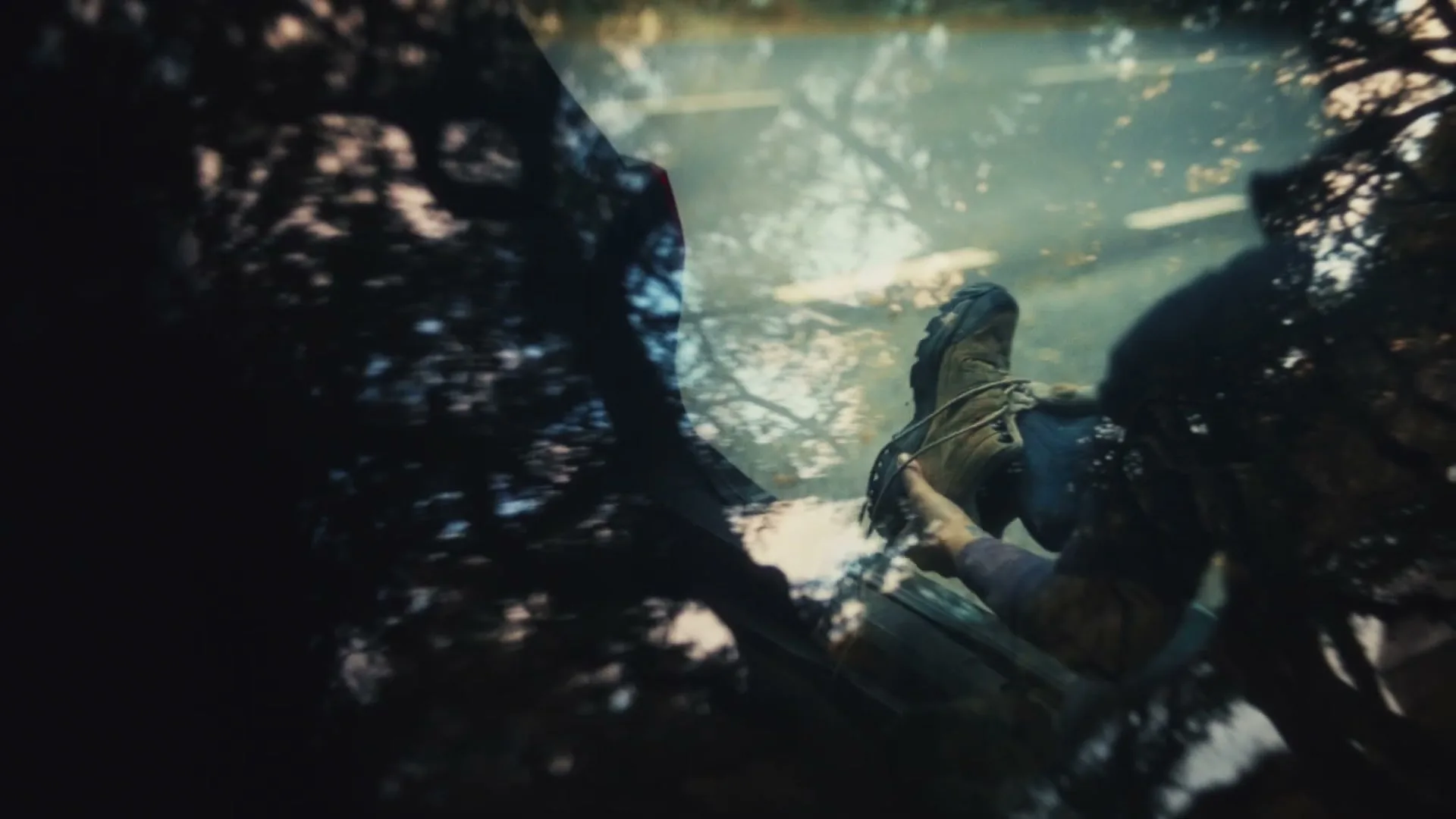 A person's hand holds a hiking boot, reflected in a puddle with trees and sky visible, suggesting a journey's end or pause.