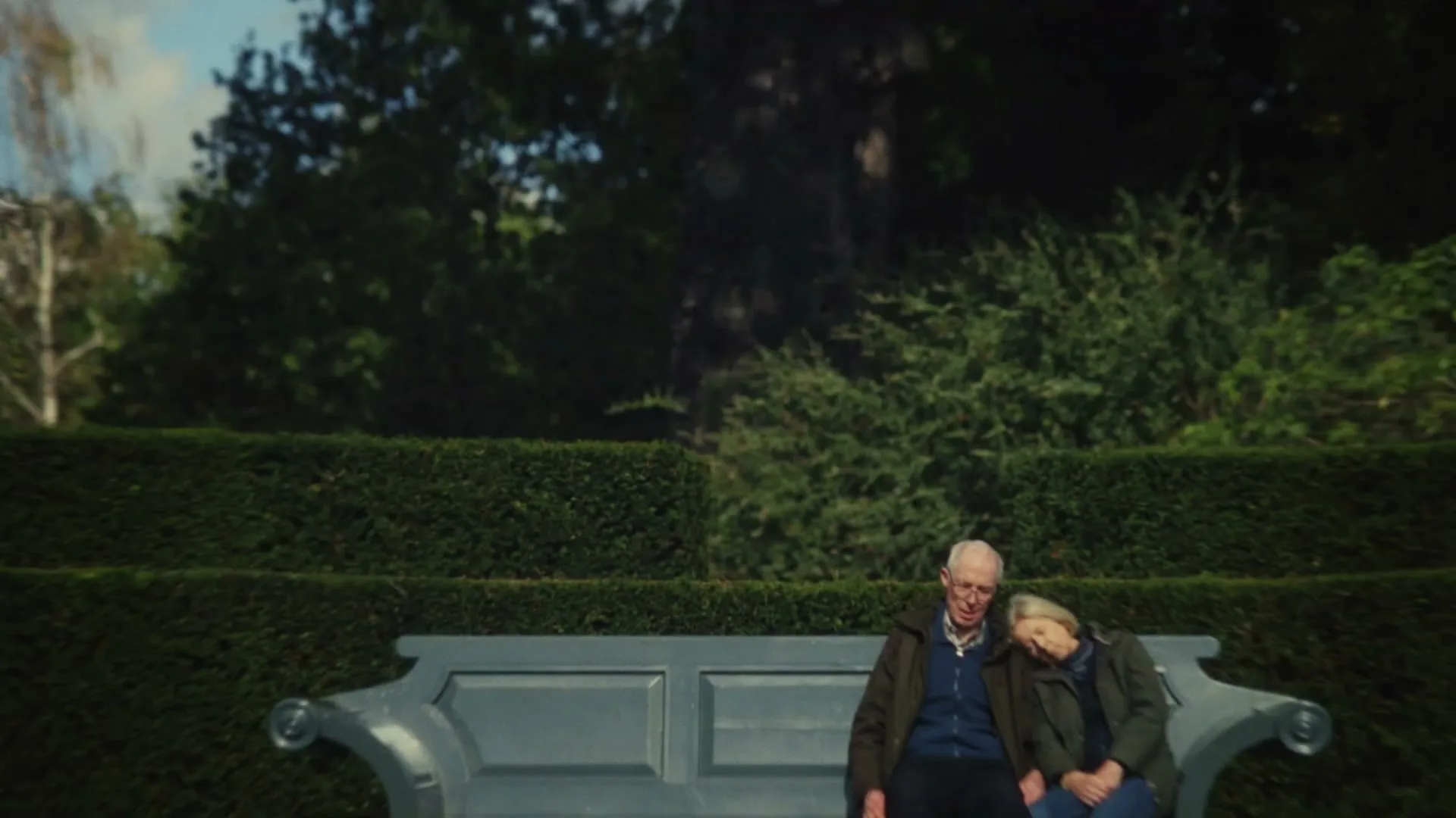A blurred blonde woman walks past an elderly couple sitting on a bench in a garden, with the woman resting her head on the man's shoulder.