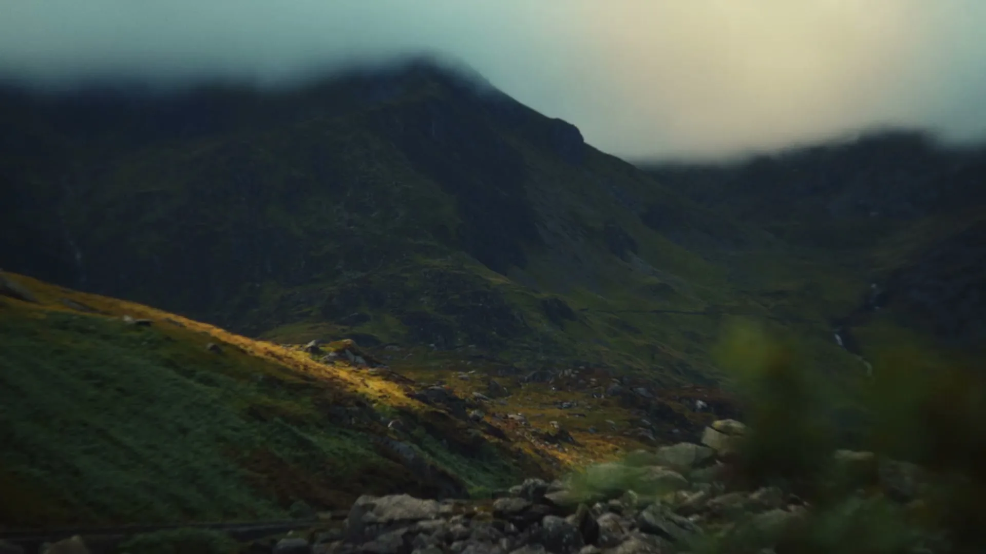 A wide shot of a vast, green mountain landscape with low-hanging clouds and a patch of sunlight illuminating the lower slopes.