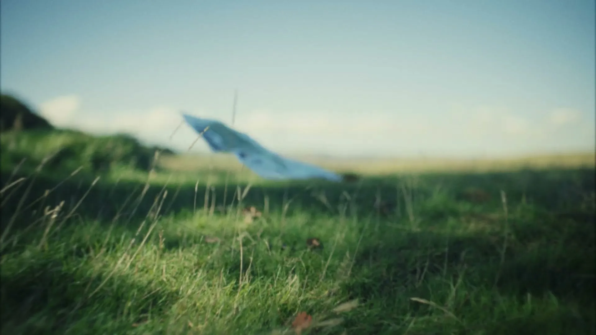 A blurred map, possibly a paper kite, flies low over a grassy field under a bright sky, with tall grass in the foreground.