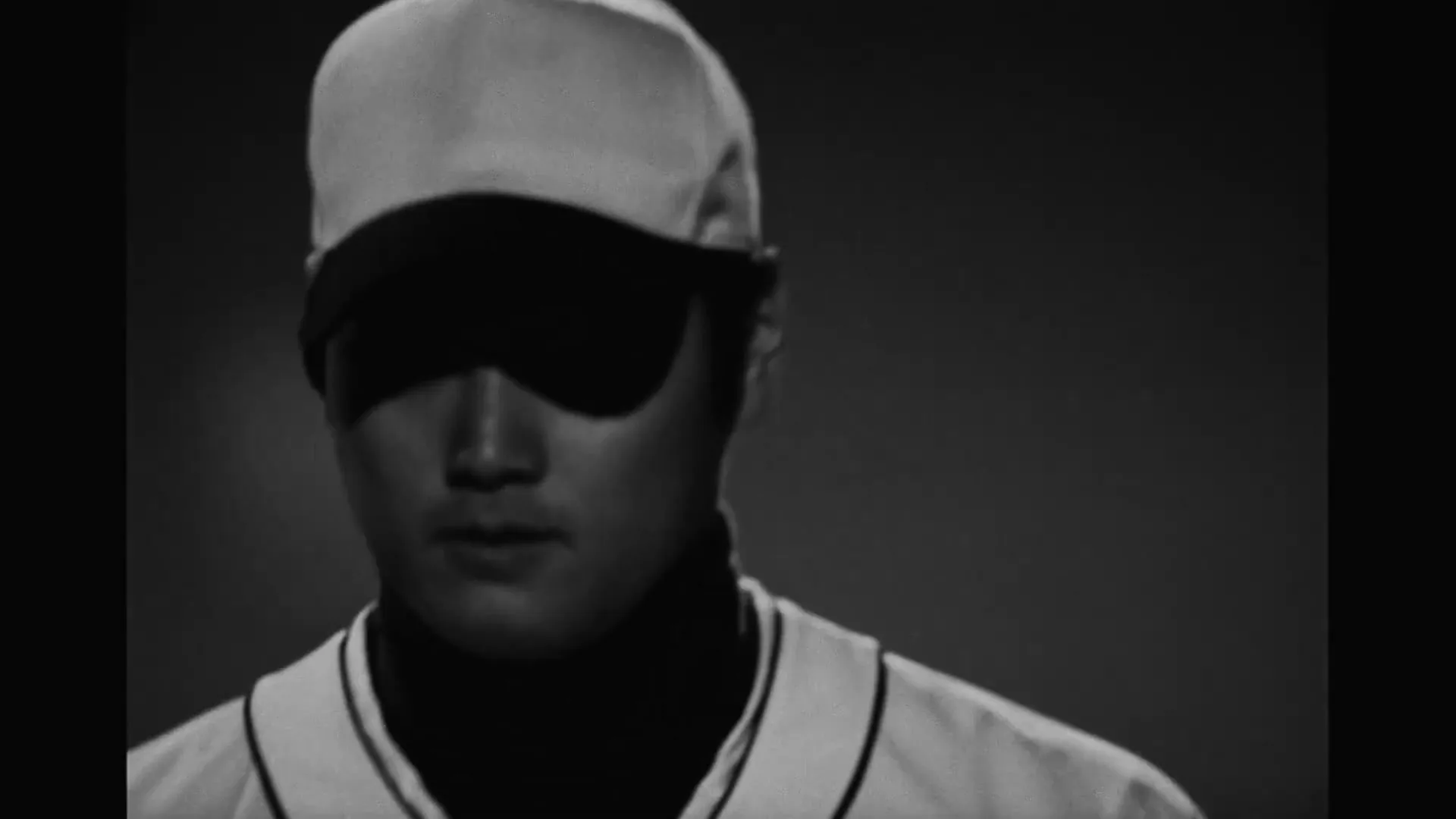 A male baseball player in a cap and uniform practices his swing in a dark, smoky batting cage, then walks away.