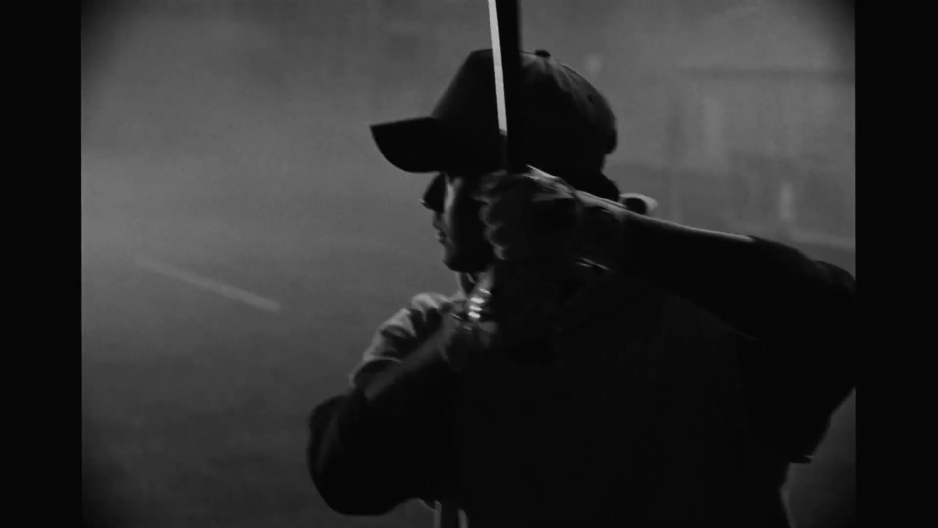 A male baseball player in a cap and hoodie holds a bat, silhouetted against a smoky background in a black and white shot.