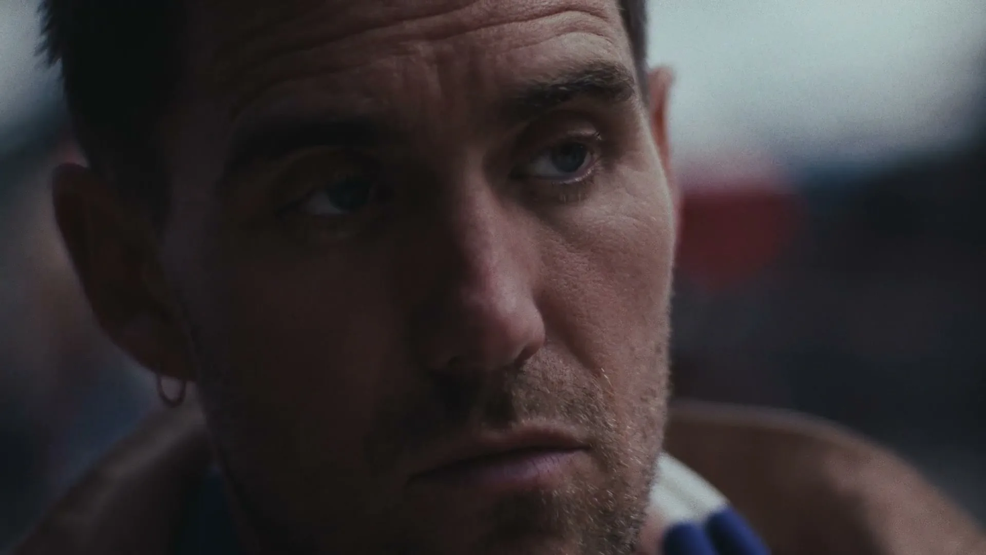 A man with short brown hair and stubble looks down, then up and to the right, with a slight smile, in a dimly lit interior.