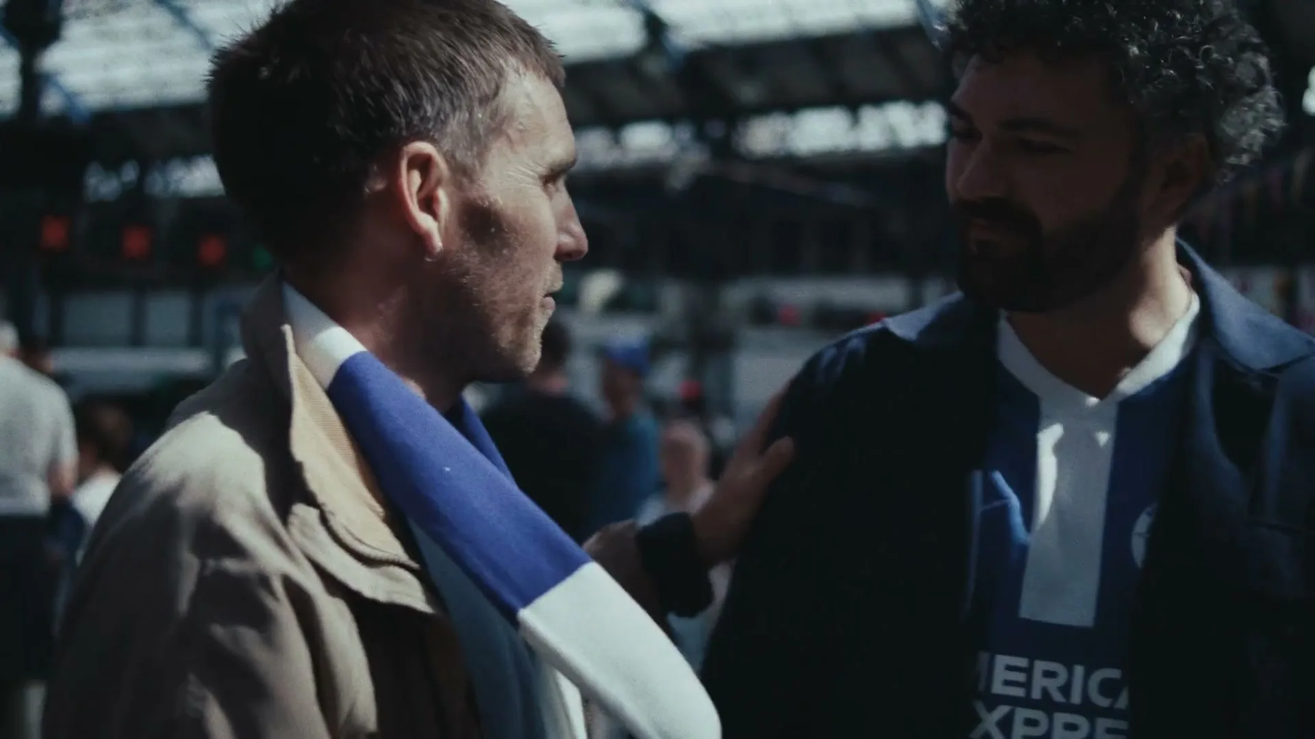 Two male football fans, one wearing a Brighton & Hove Albion shirt, talk and walk through a busy train station.