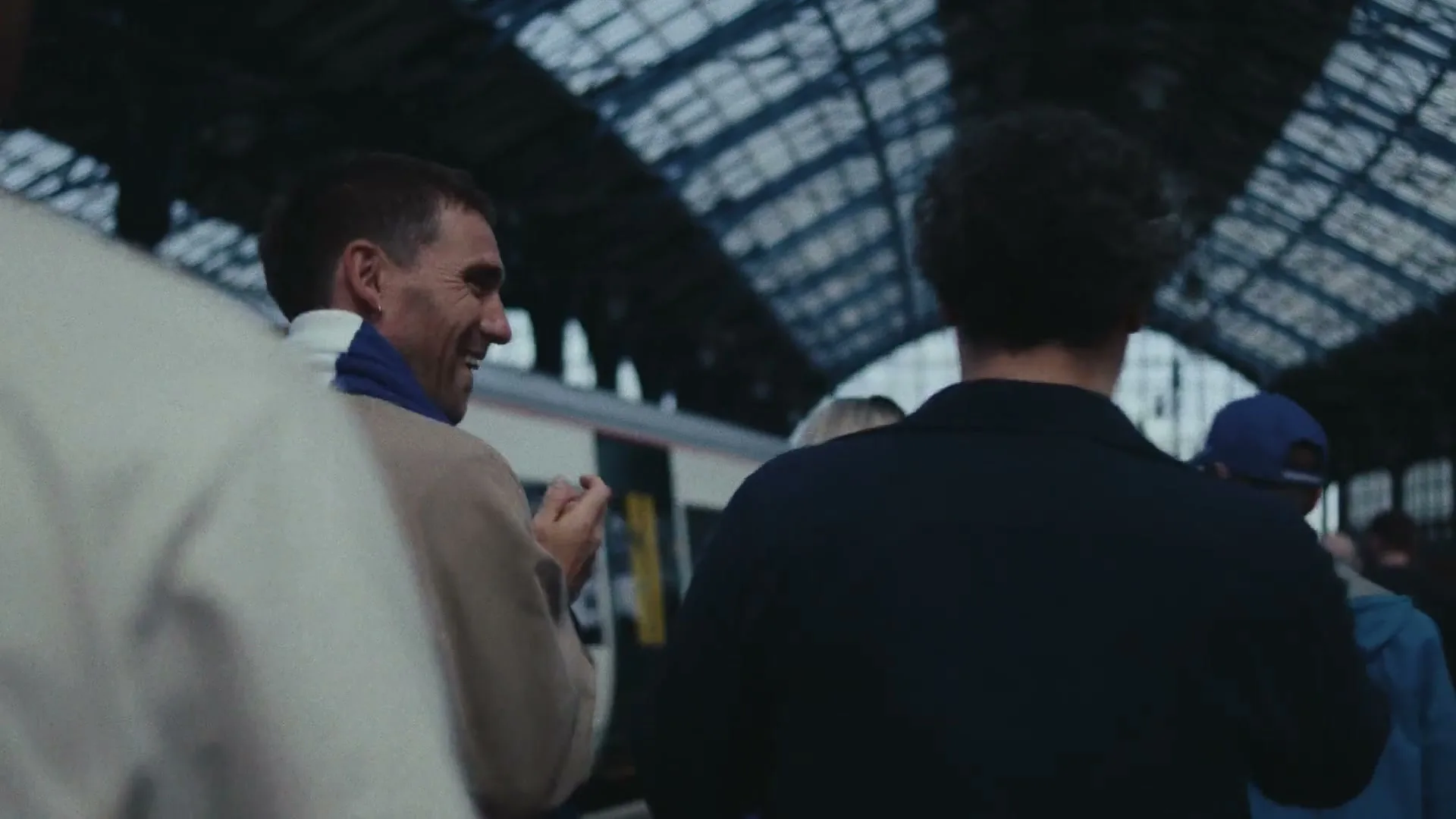 A man in a beige jacket and blue scarf smiles and claps while standing on a train platform with other people.