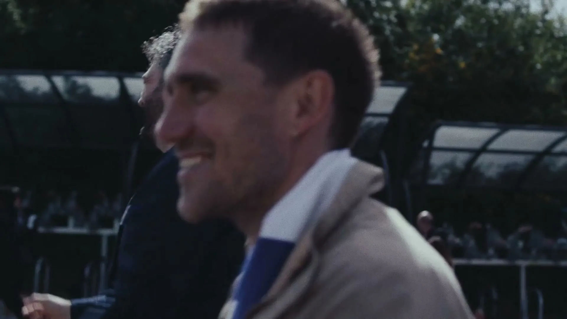 Two men, one with a beard and one with short hair, walk and smile in an outdoor stadium tunnel on a bright day.
