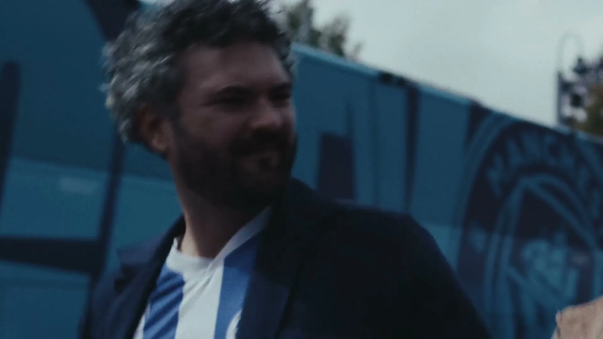 A man with curly hair and a beard, wearing a dark jacket over a blue and white striped shirt, walks past a blue wall with a logo.