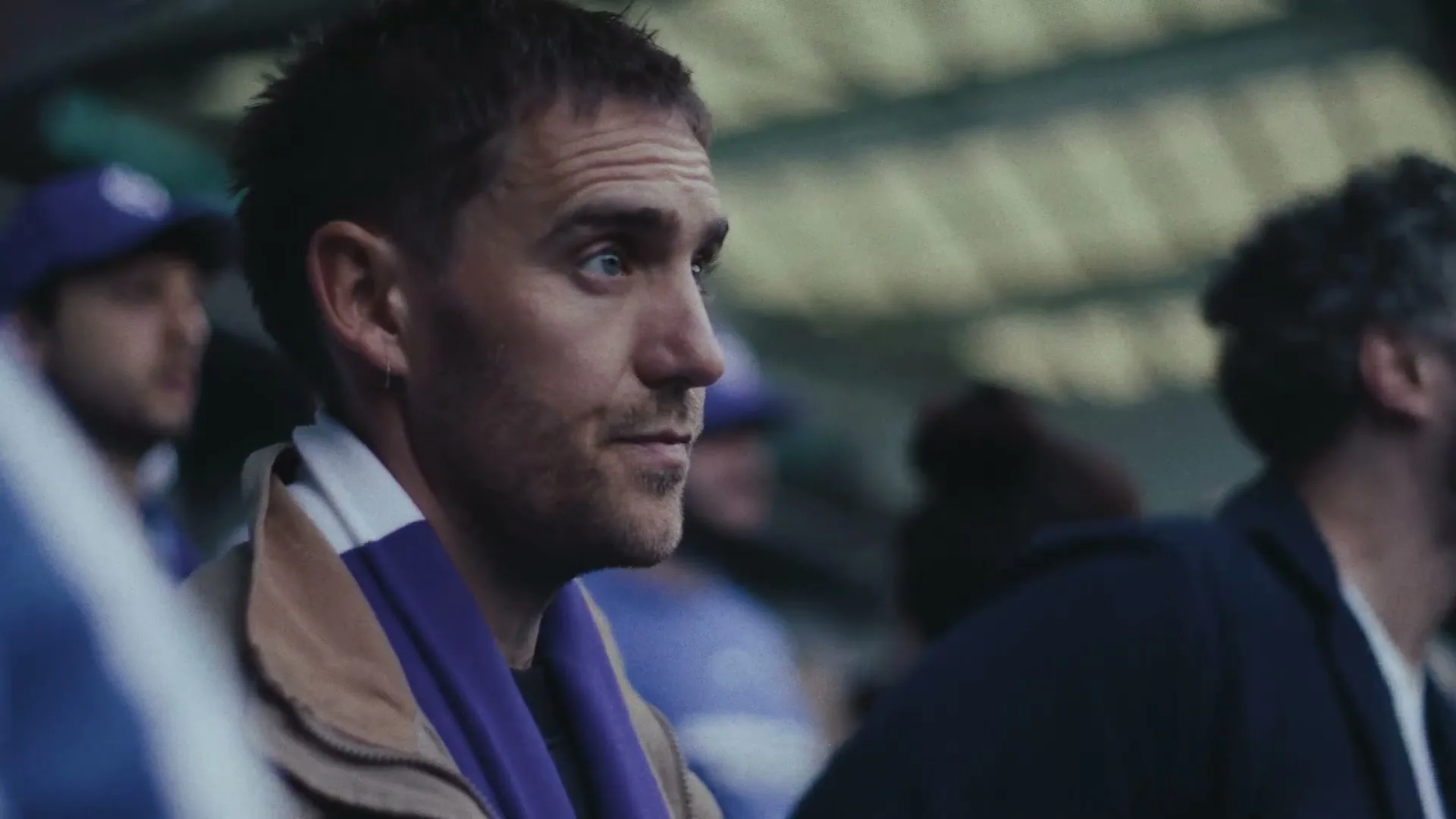 A male football fan in his 30s, wearing a brown jacket and purple scarf, watches the game intently from the stands of a stadium.
