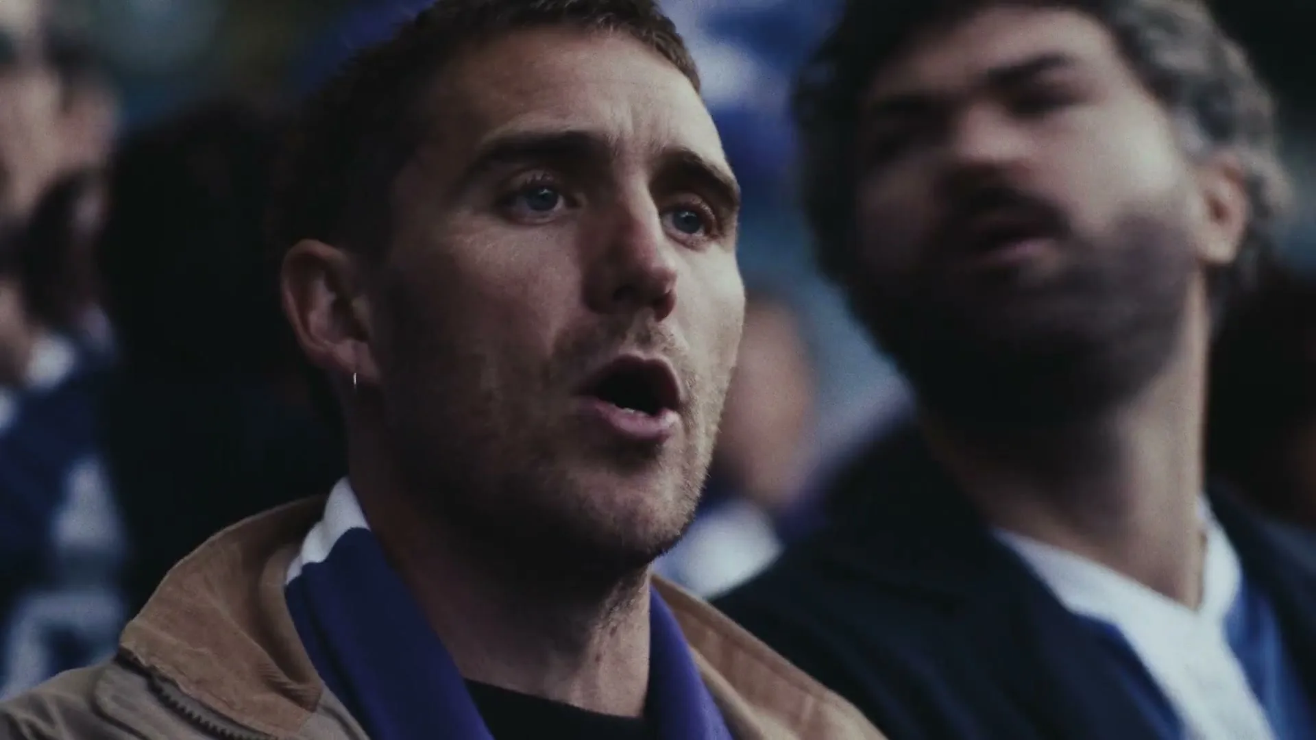 Two male football fans, one with a beard, watch a game intently in a stadium, wearing team scarves.