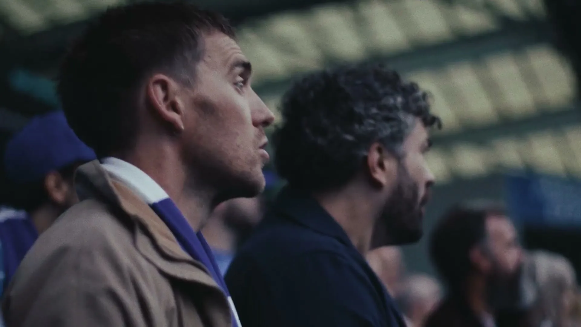 Two men, one with short brown hair and a tan jacket, the other with curly grey hair, watch a football match from the stands.