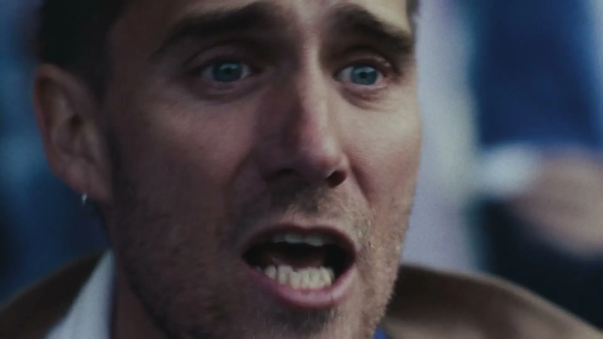 A man with blue eyes and stubble, wearing a brown jacket, looks distressed and whispers, his mouth open, in a blurry, dark setting.