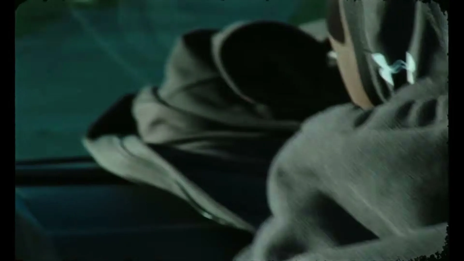 A person in a dark hoodie with an Under Armour logo is seen from the side, getting out of a car in a dark, moody setting.