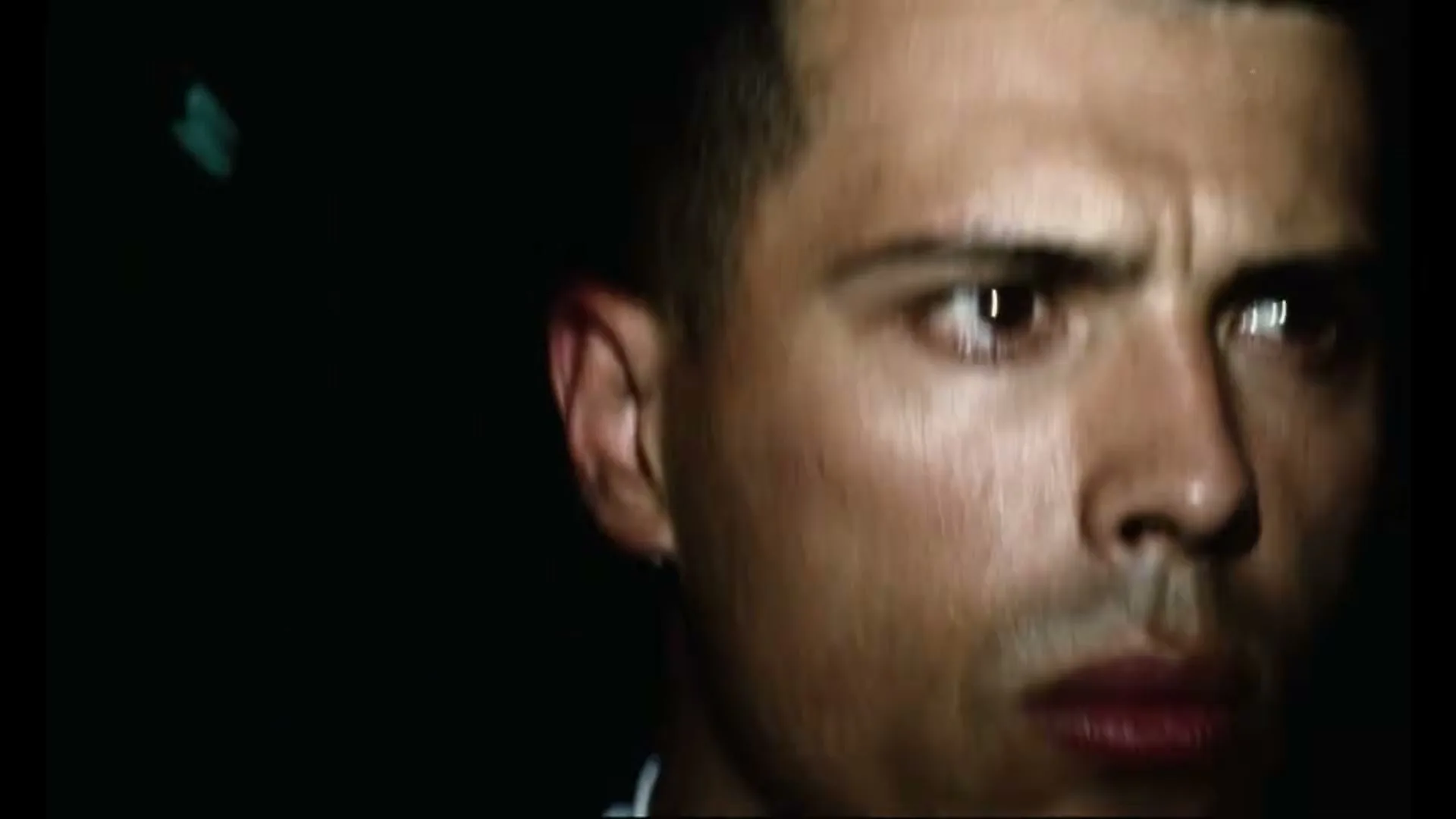 A male athlete in his 20s, wearing a white compression shirt, looks intensely forward in a dark, indeterminate setting.
