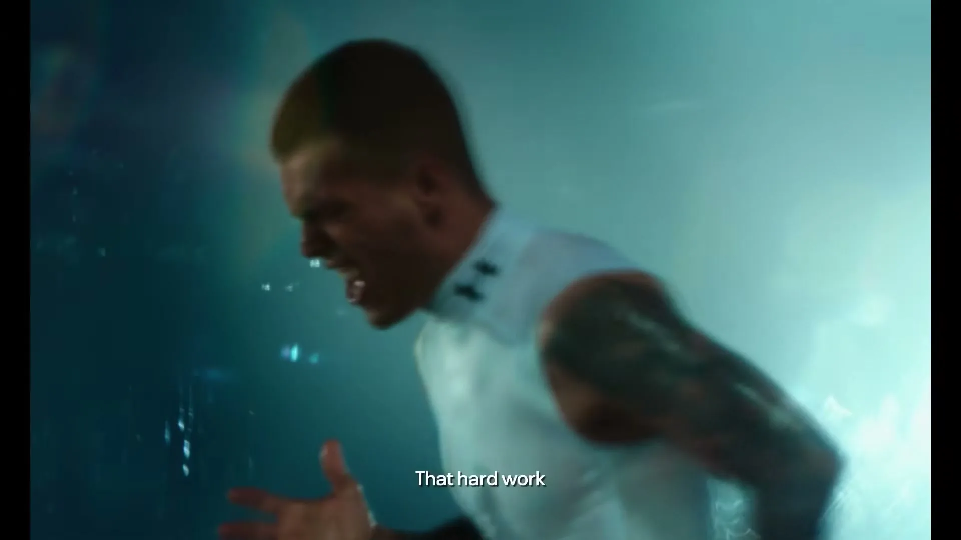A male athlete in a white compression shirt with tattoos on his arm, screams in a dark, misty, blue-lit environment.