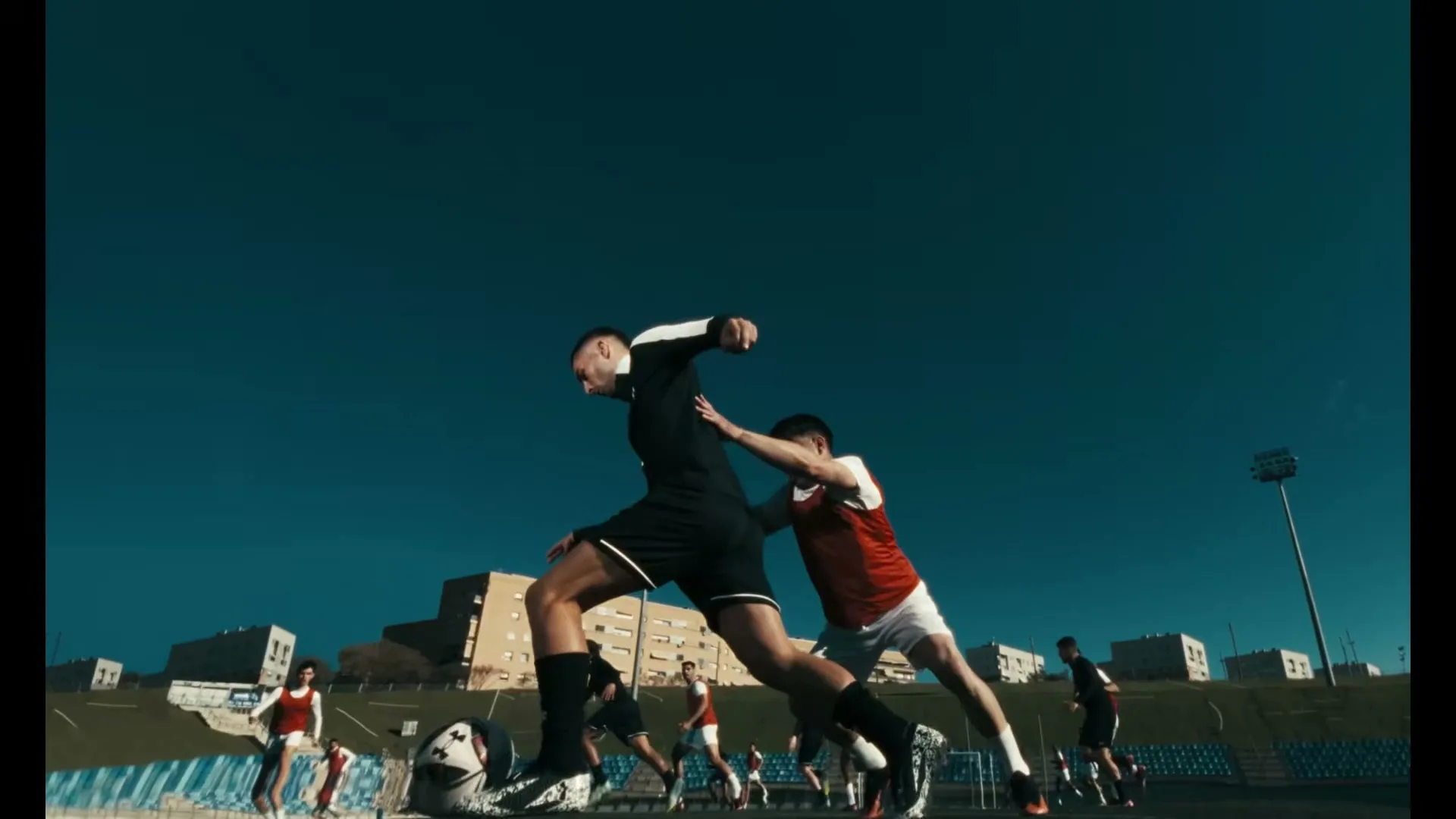 Two male soccer players, one in black and one in red, struggle for the ball on a sunny outdoor field with buildings in the background.