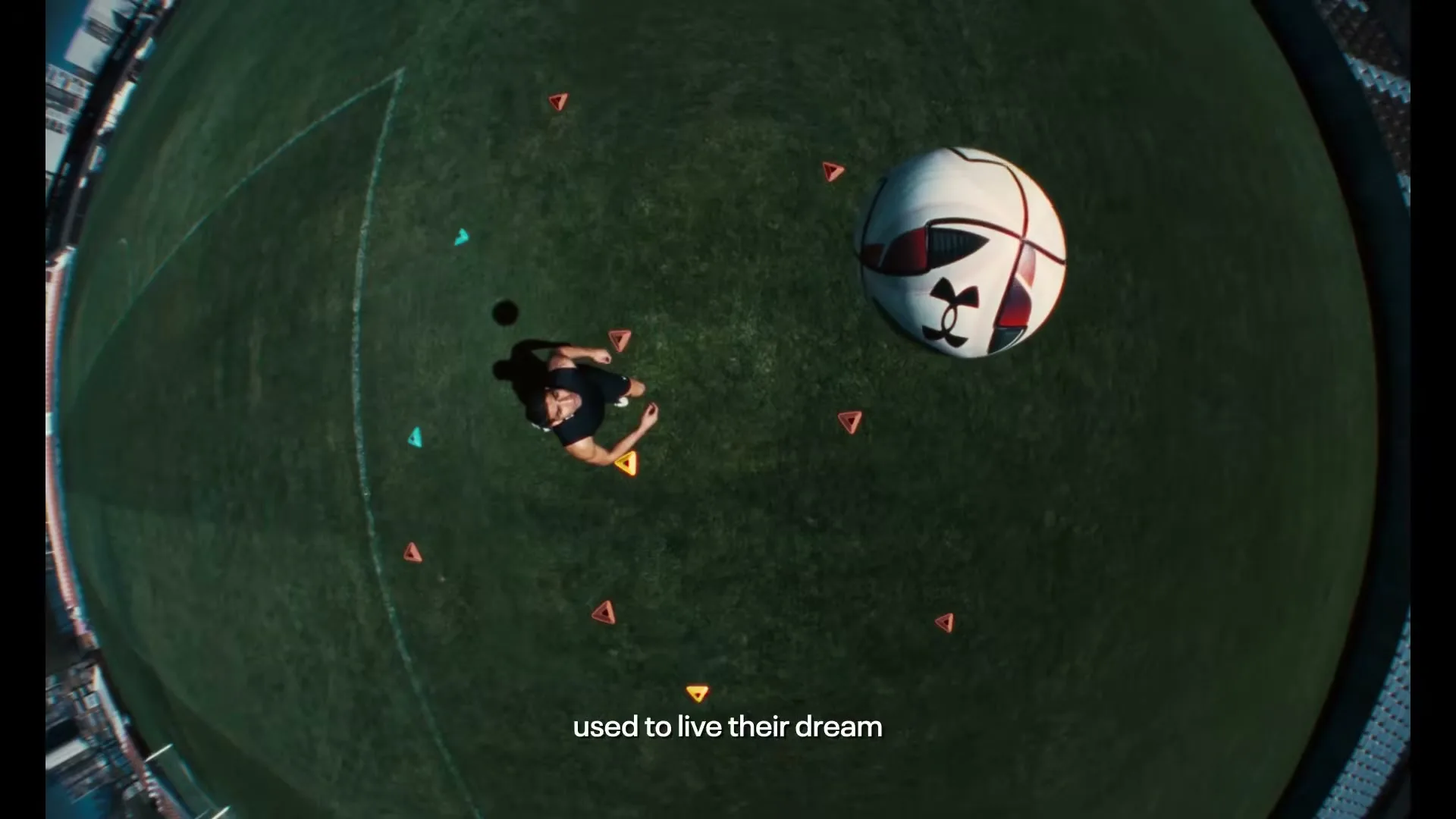 A male soccer player in black athletic wear looks up at a large soccer ball floating above him on a green field with orange cones.