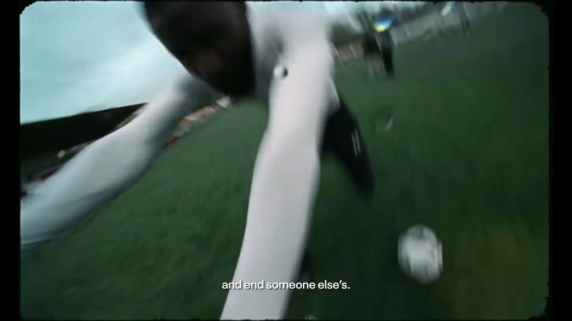 Two male athletes in white and black uniforms tackle each other on a green football field under a cloudy sky, with other players in the background.
