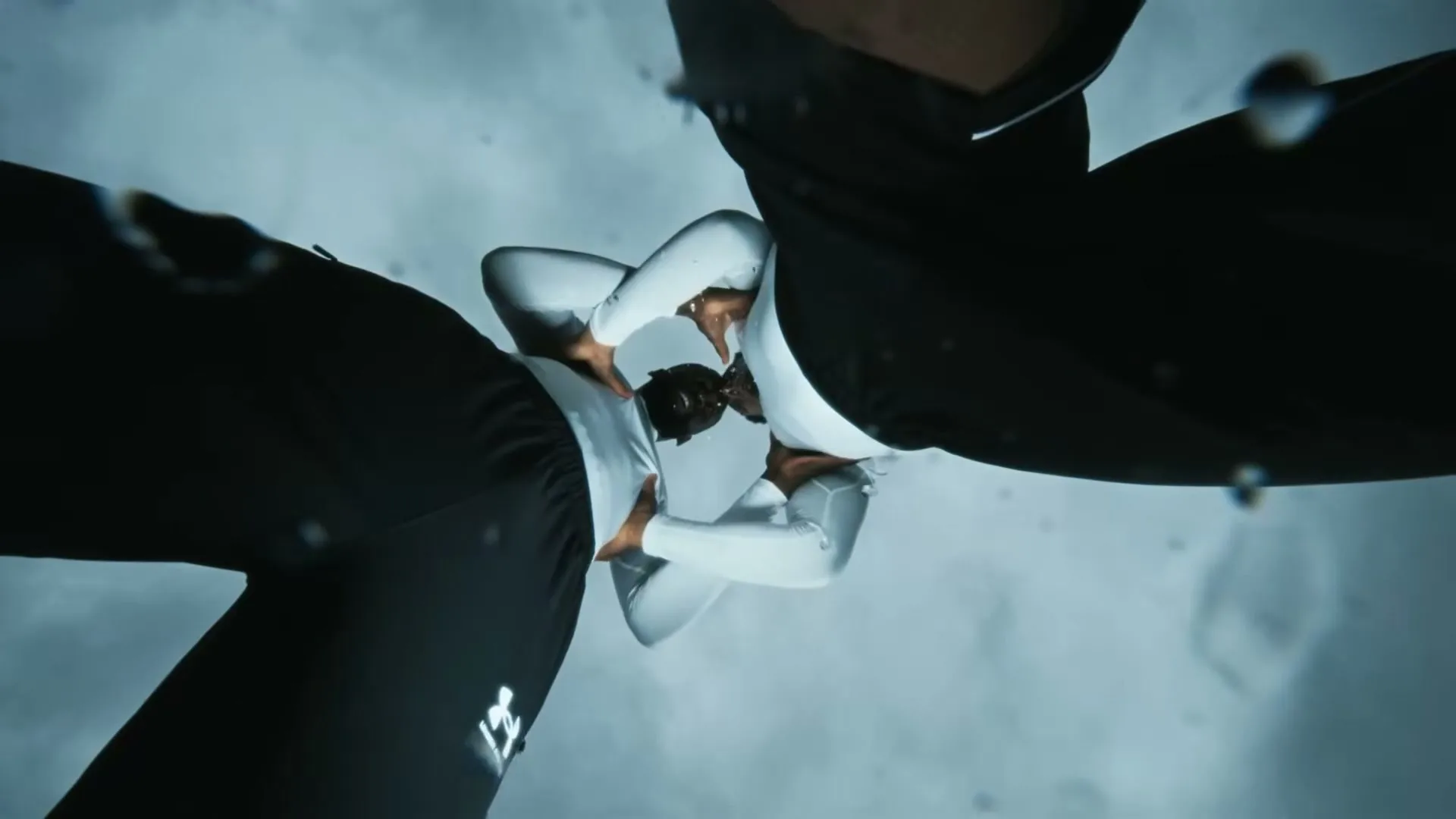 A male athlete in black and white sportswear is seen from an extreme low angle, looking up at the camera as water droplets fall around him.