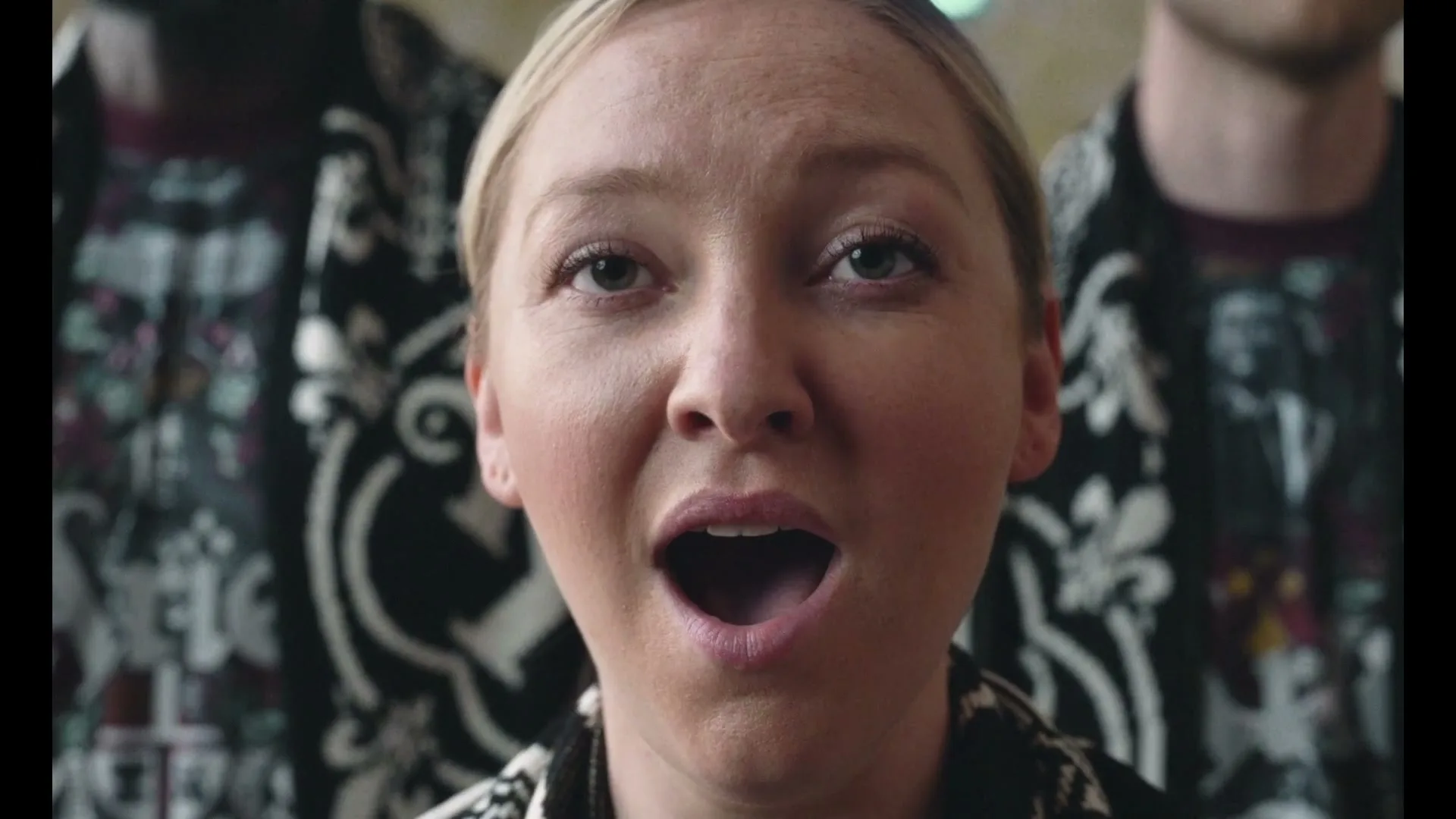 A blonde woman in her 20s, wearing a patterned shirt, sings with her mouth open, looking up in an interior setting.