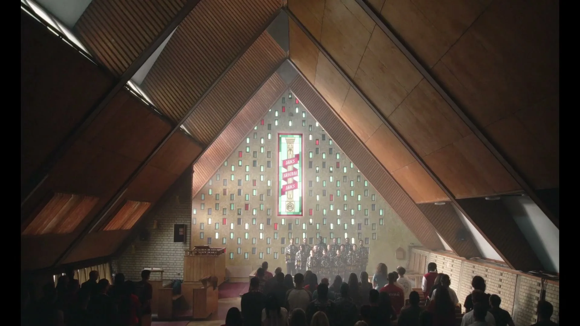 A wide shot from a high angle shows a diverse congregation in a modern church, with a choir performing in front of a stained-glass window.
