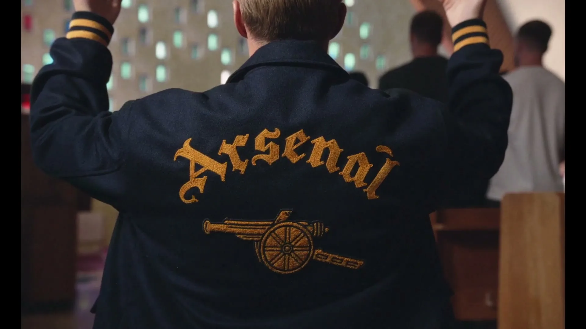 A man with blonde hair, wearing a dark blue jacket with "Arsenal" and a cannon logo on the back, stands with his arms raised in a church.
