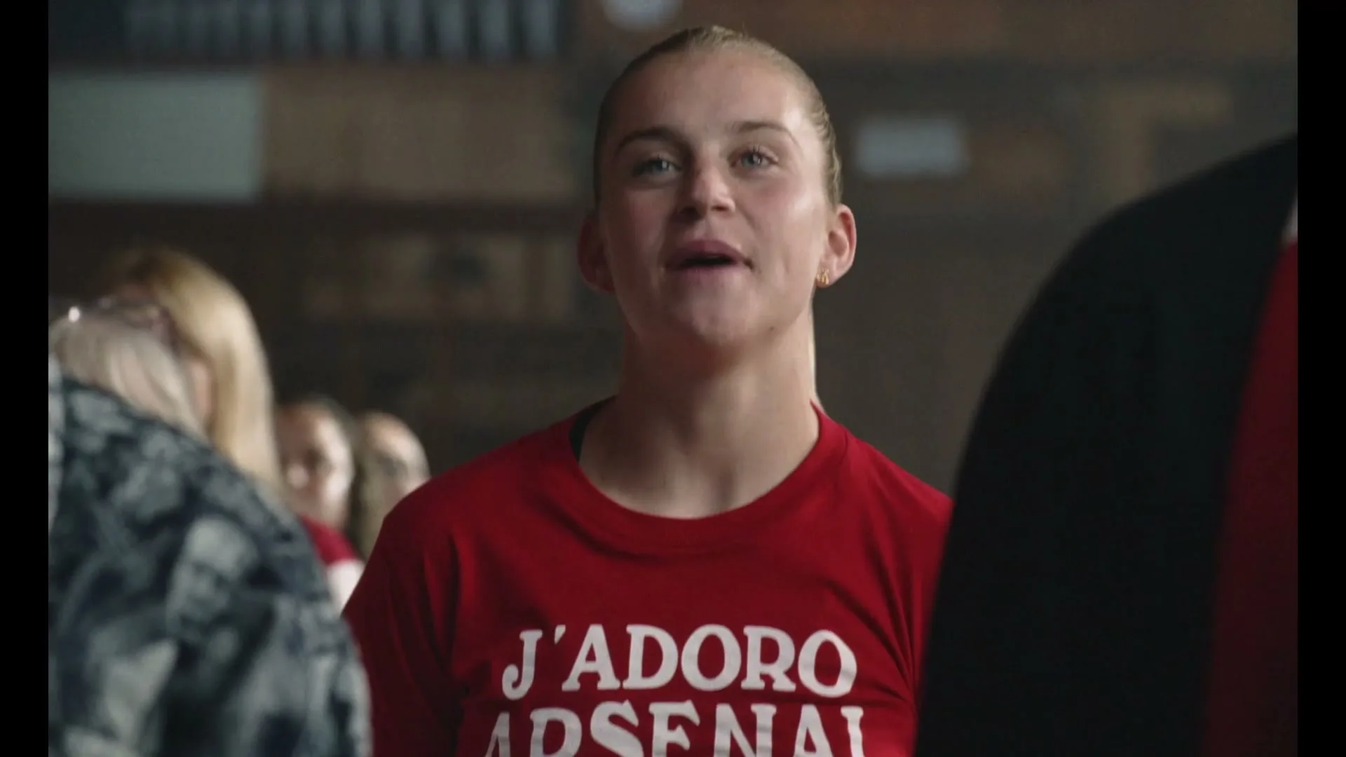 A young woman in a red t-shirt with "J'ADORE ARSENAL" printed on it looks up and smiles, surrounded by other people in a large, dimly lit room.