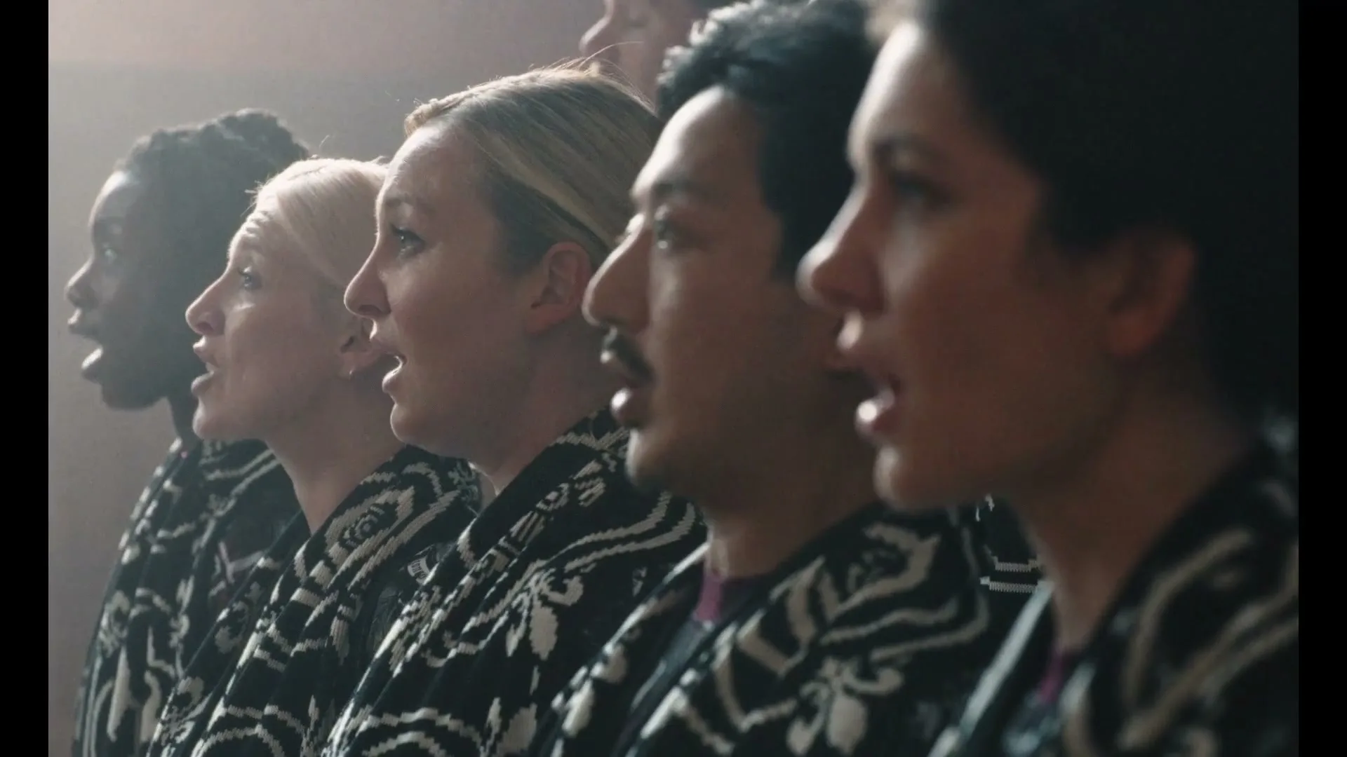 A diverse group of people, dressed in matching black and white patterned robes, sing in unison, their mouths open wide, in a dimly lit interior.