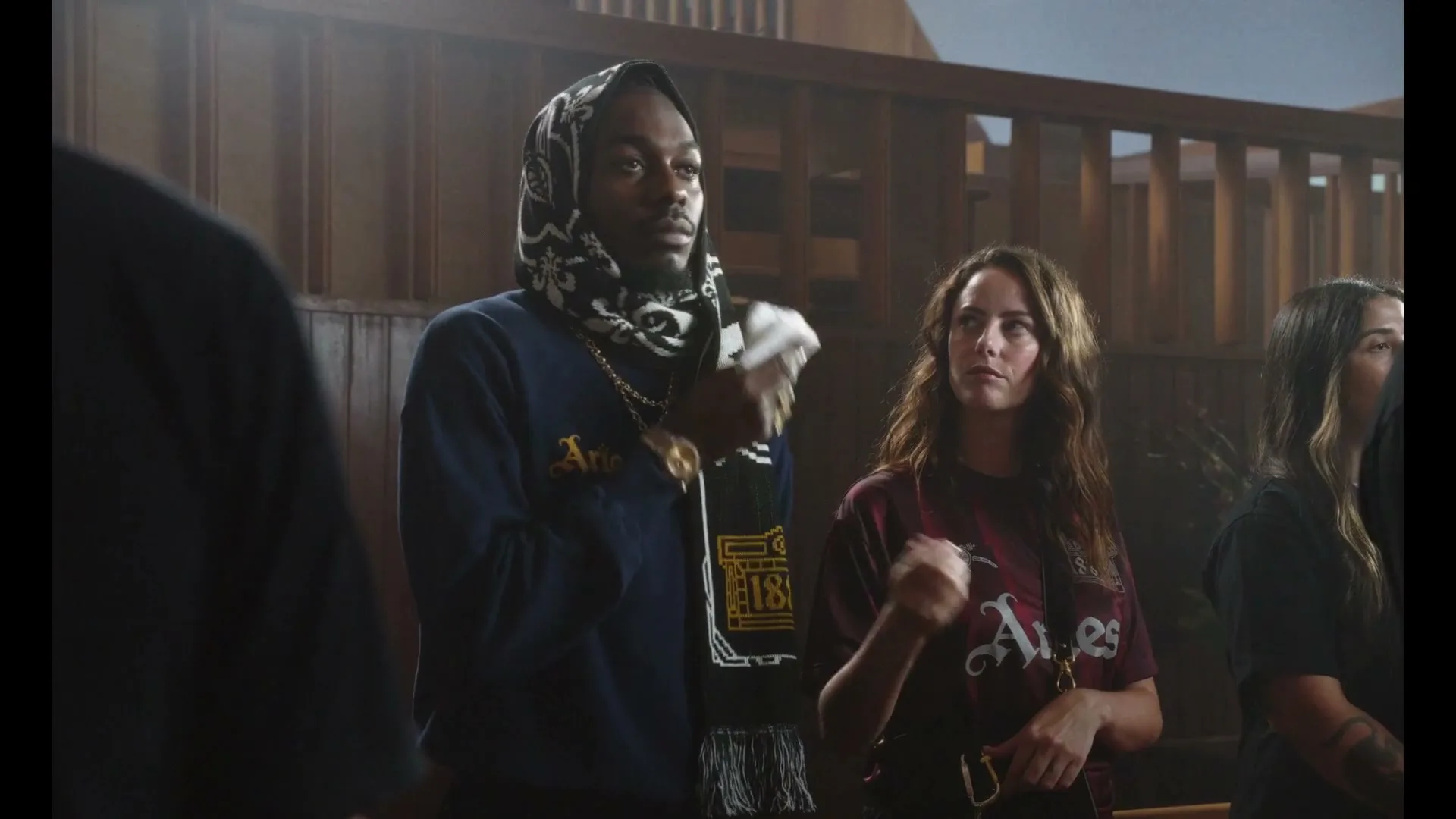 A young man in a dark blue sweatshirt and a black and white scarf over his head stands with a woman in a maroon t-shirt, both looking up in a wooden room.