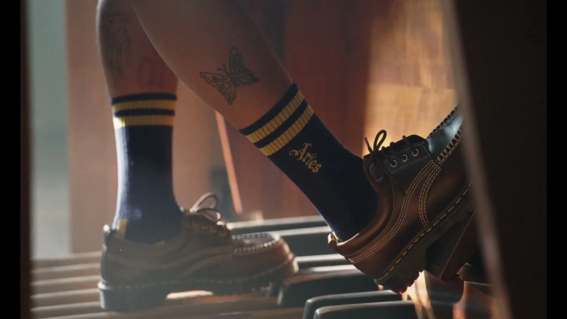 A person's legs with tattoos and blue socks with yellow stripes and "Aries" written on them, wearing brown shoes, playing an organ.