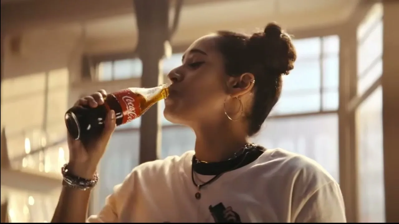A young woman with dark hair in a bun drinks from a glass bottle of Coca-Cola, then smiles, in an industrial-style interior.