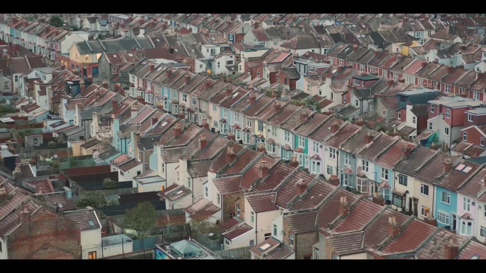 An aerial view shows rows of colorful terraced houses with red roofs stretching into the distance under an overcast sky.