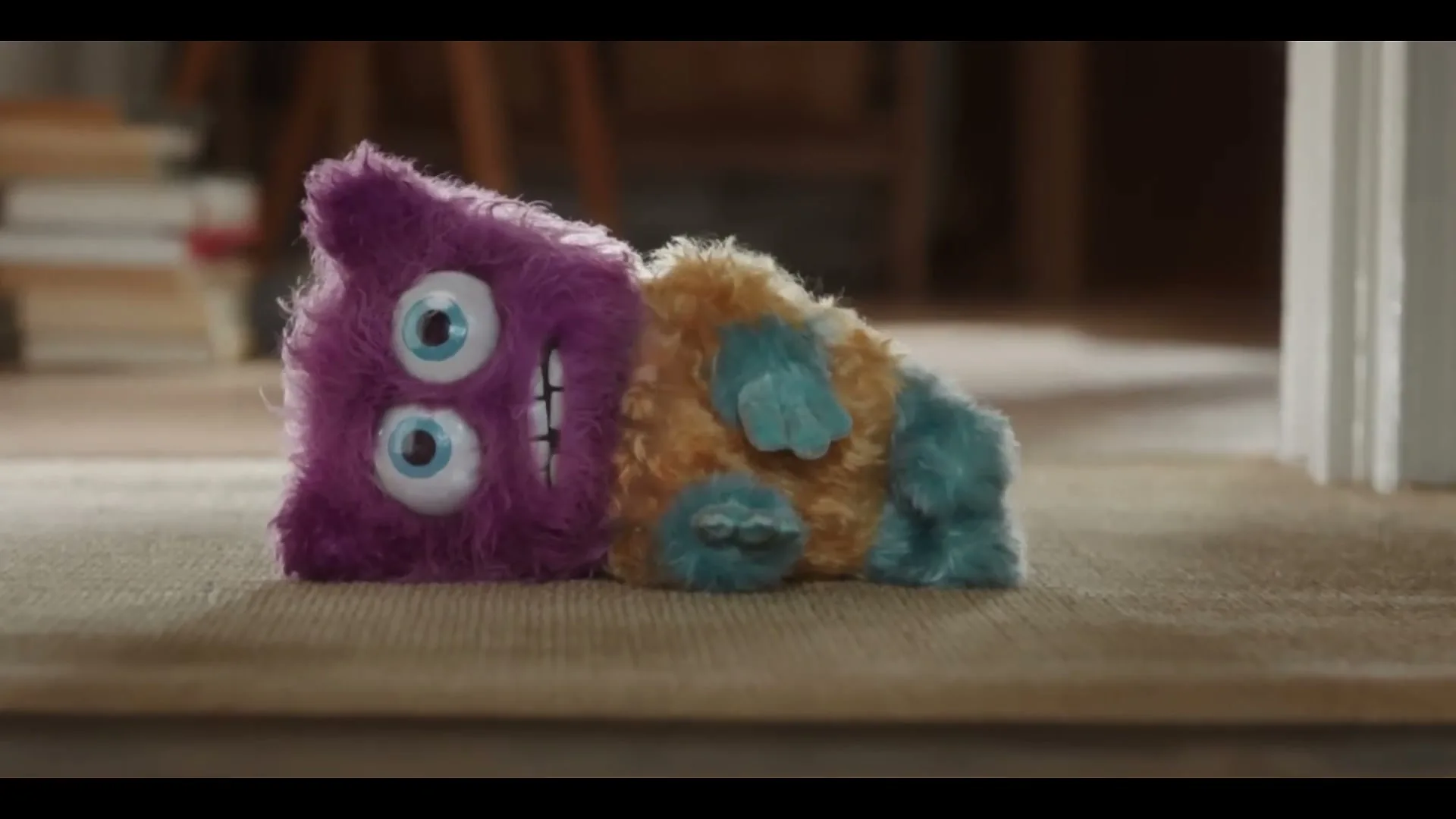 A purple and orange furry monster toy with big eyes and a wide smile lies on a carpeted floor in a home.
