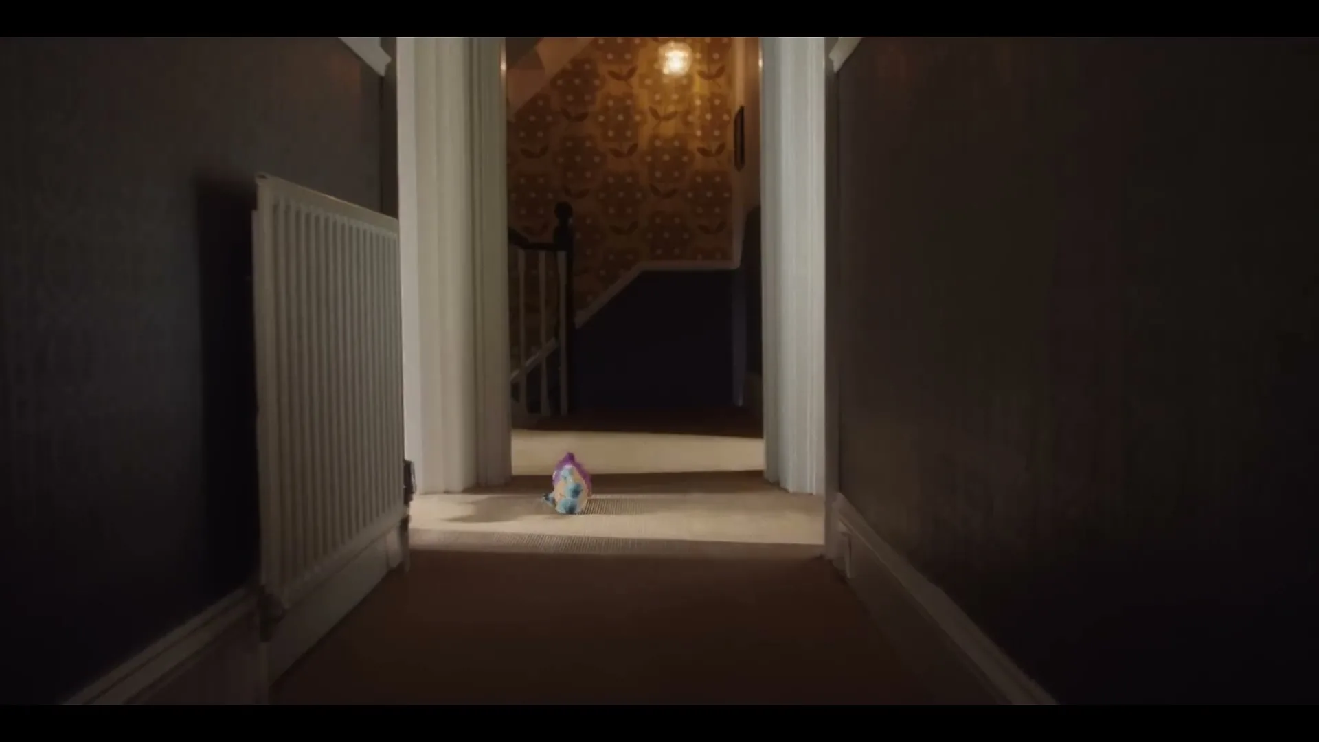 A small, colorful toy unicorn sits alone on the carpeted floor of a dimly lit, narrow hallway in a house.