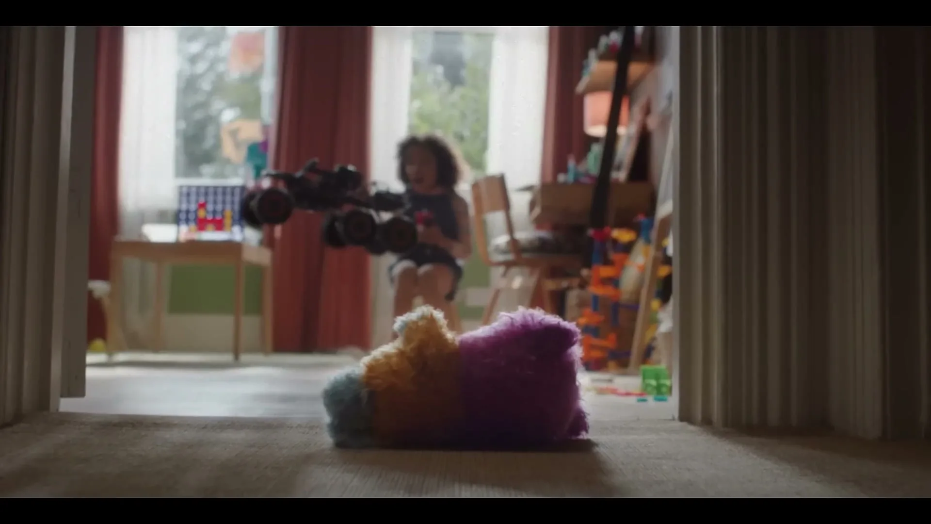 A child plays with a remote control car, making it fly over a colorful plush toy on the floor of a messy bedroom.