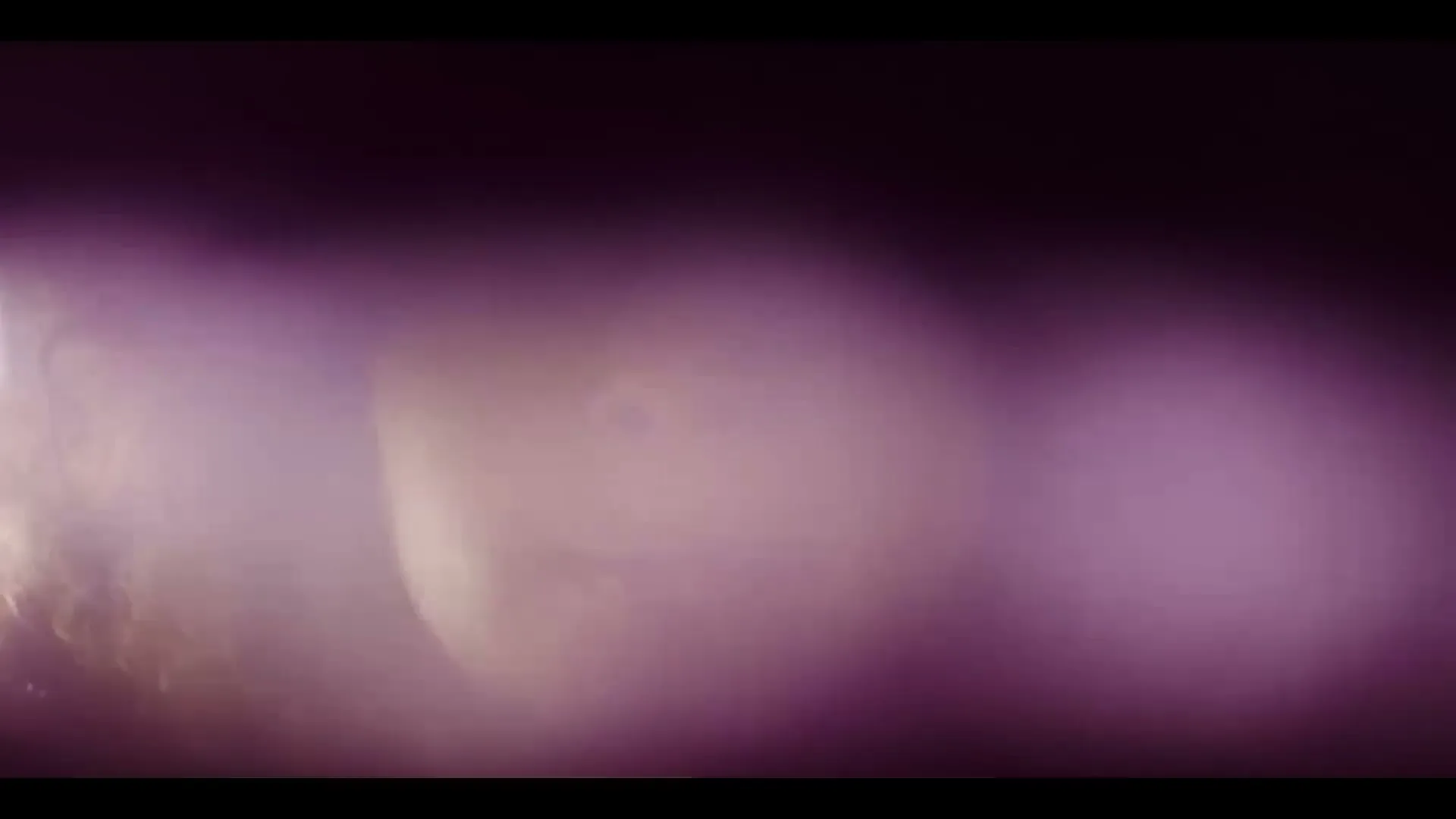 A young woman with curly hair looks directly at the camera, partially obscured by a purple lens flare.